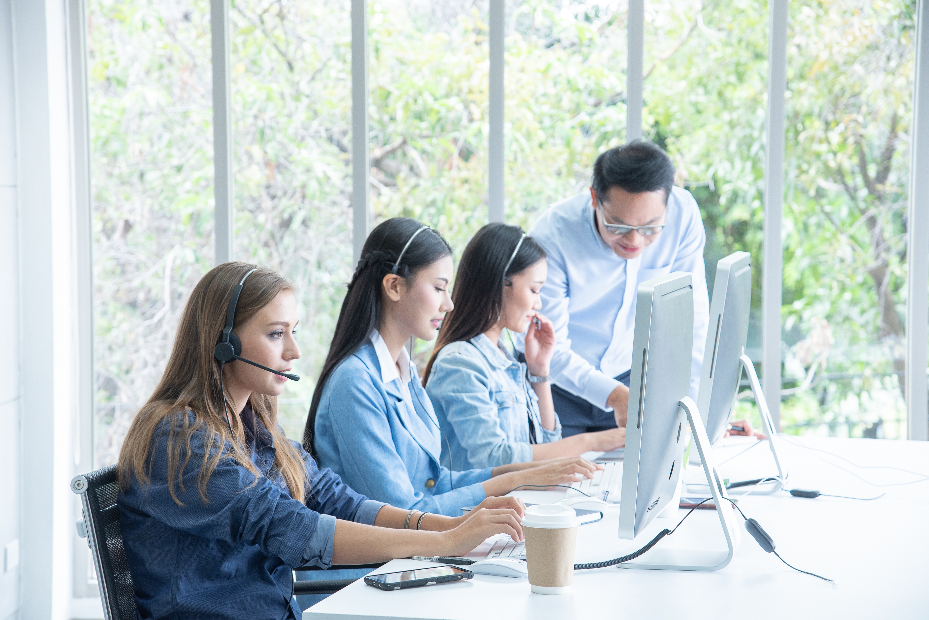 A group of call agents sitting in front of their computers. Three women are staring at their computer screen and talking into their headsets while a man is instructing the woman furthest in the picture.