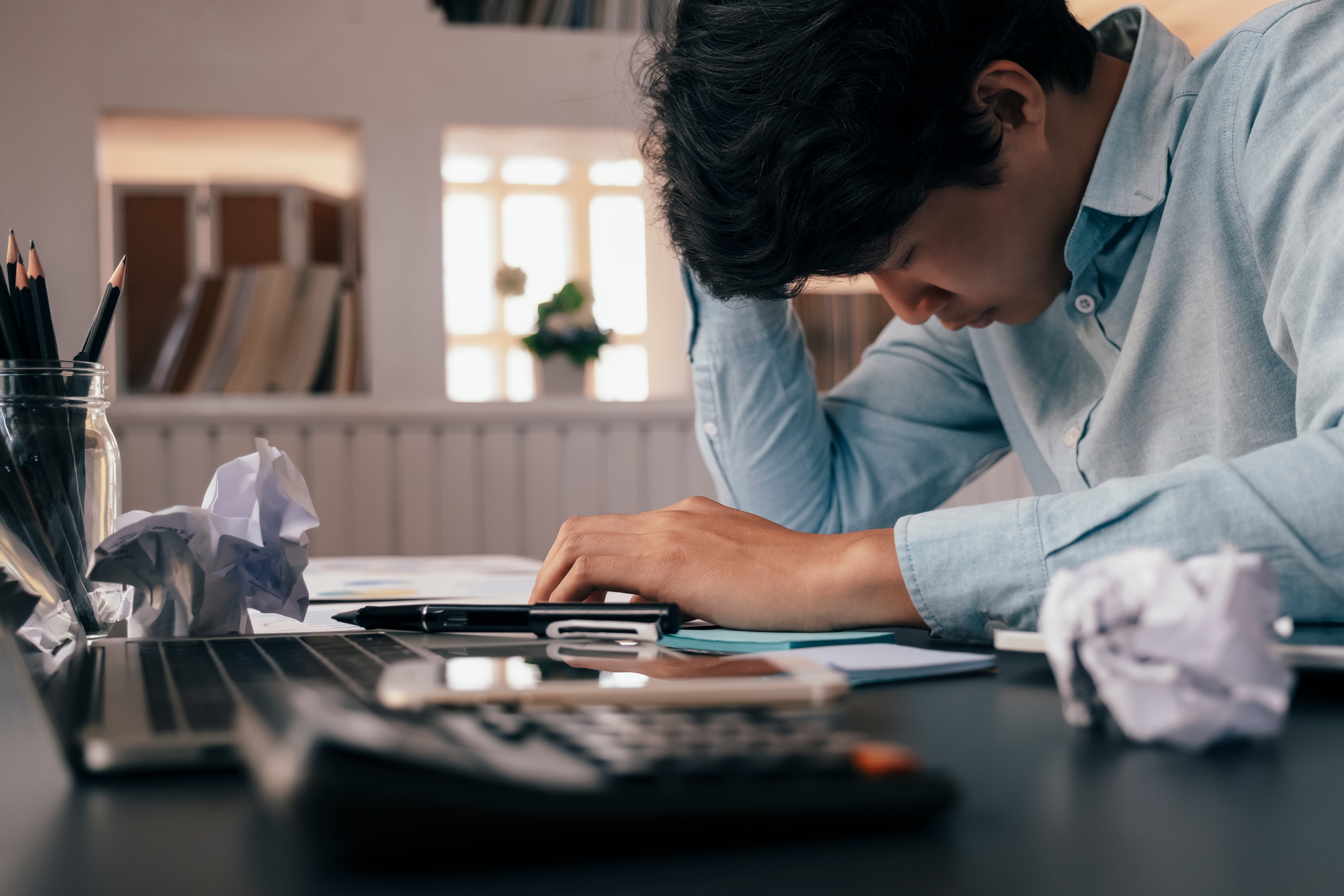 A frustrated man sitting at his desk with his head in his hand. His desk has two crumpled up pieces of paper, a notepad, a calculator, a laptop, a pen, and a bunch of sticky notes.