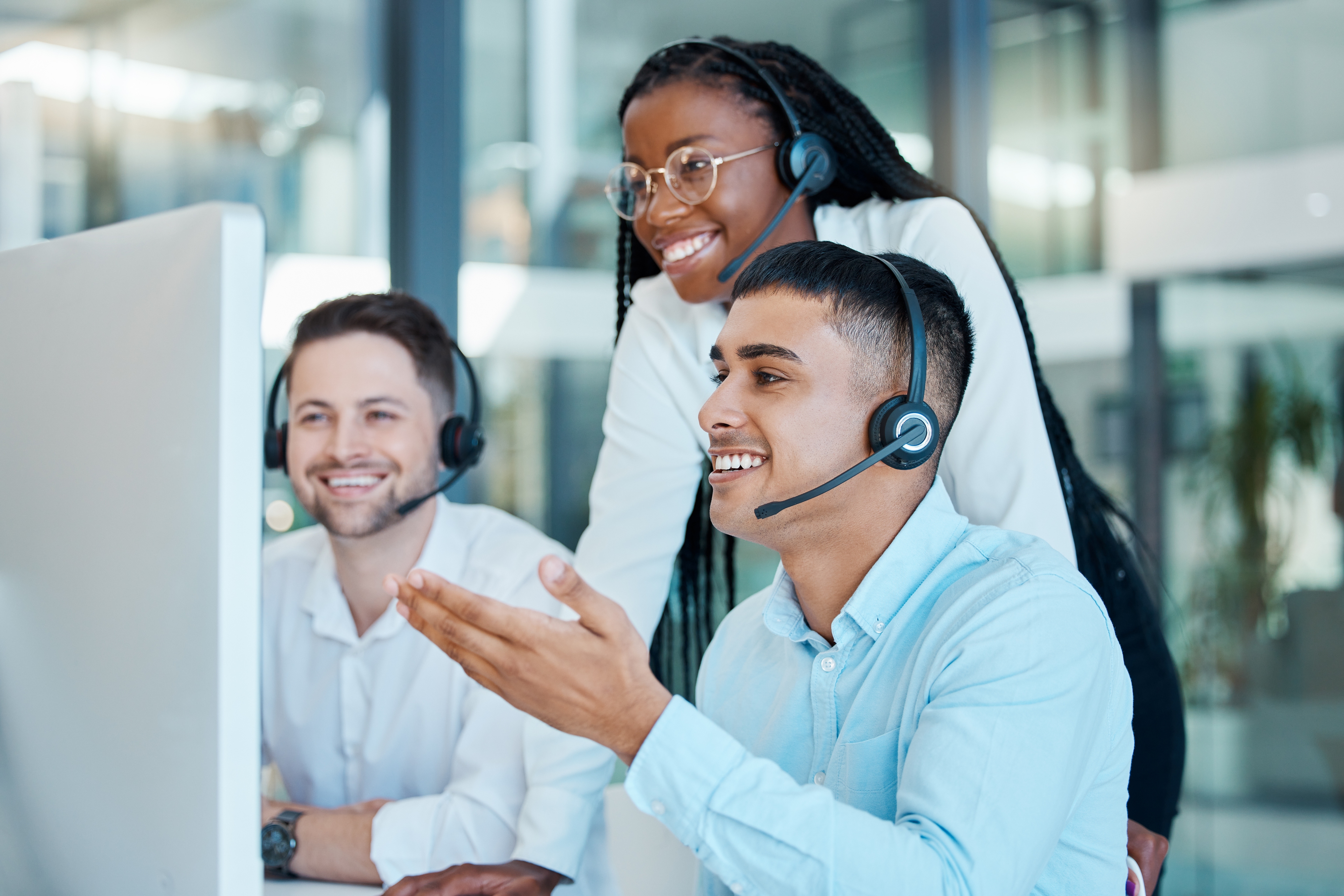 A woman training two men at a telemarketing office. They are all smiling at the computer monitor. The men are sitting at the desk while the woman looks over their shoulders.
