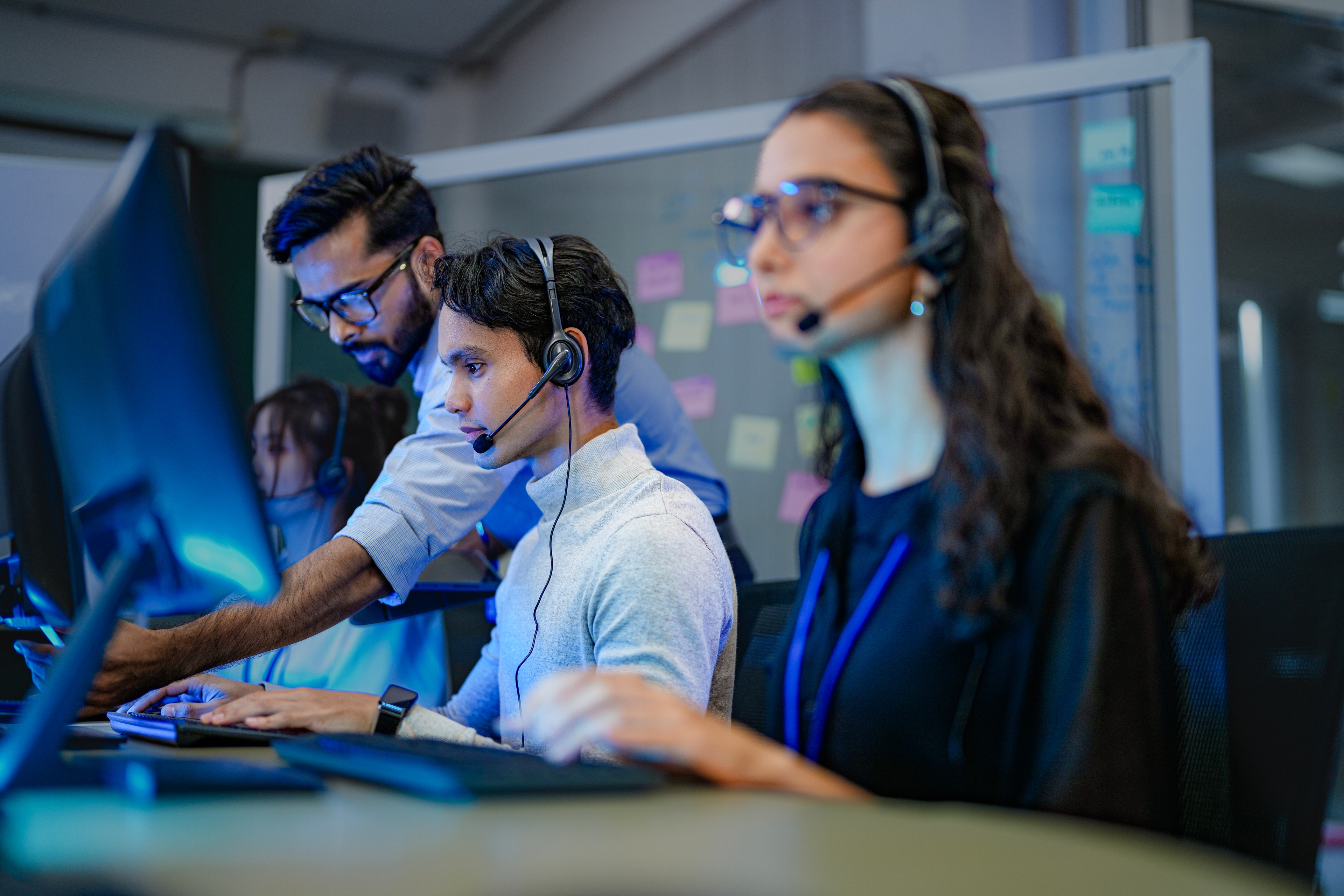 A group of call agents staring at their computers with headsets on. A man is standing over one of them and taking a look at their monitor.