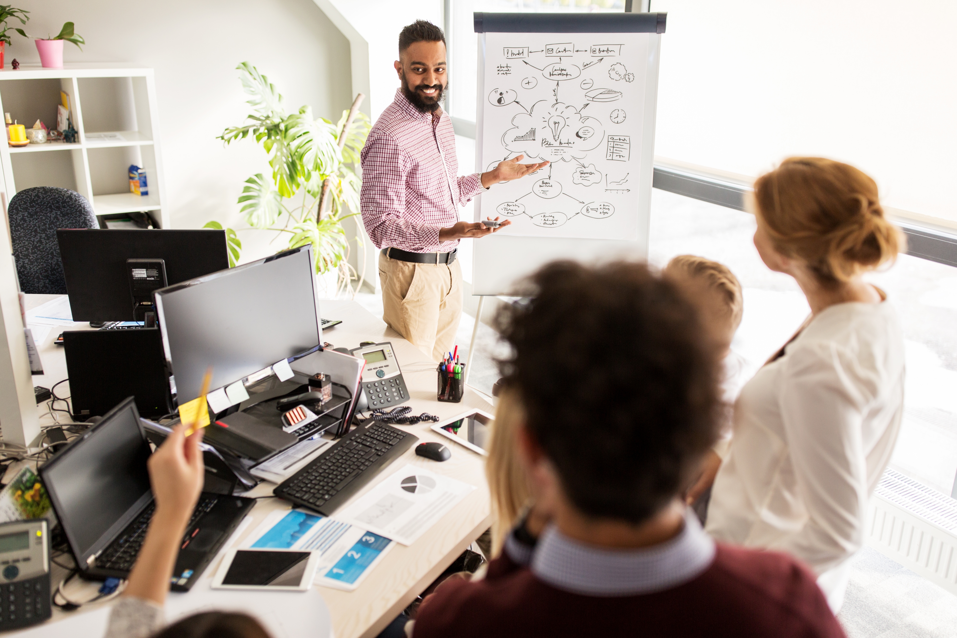 A man giving a presentation to a group of coworkers. He is gesturing to a large notepad that shows a flow of ideas. A coworker in the foreground is raising a hand with a pencil.