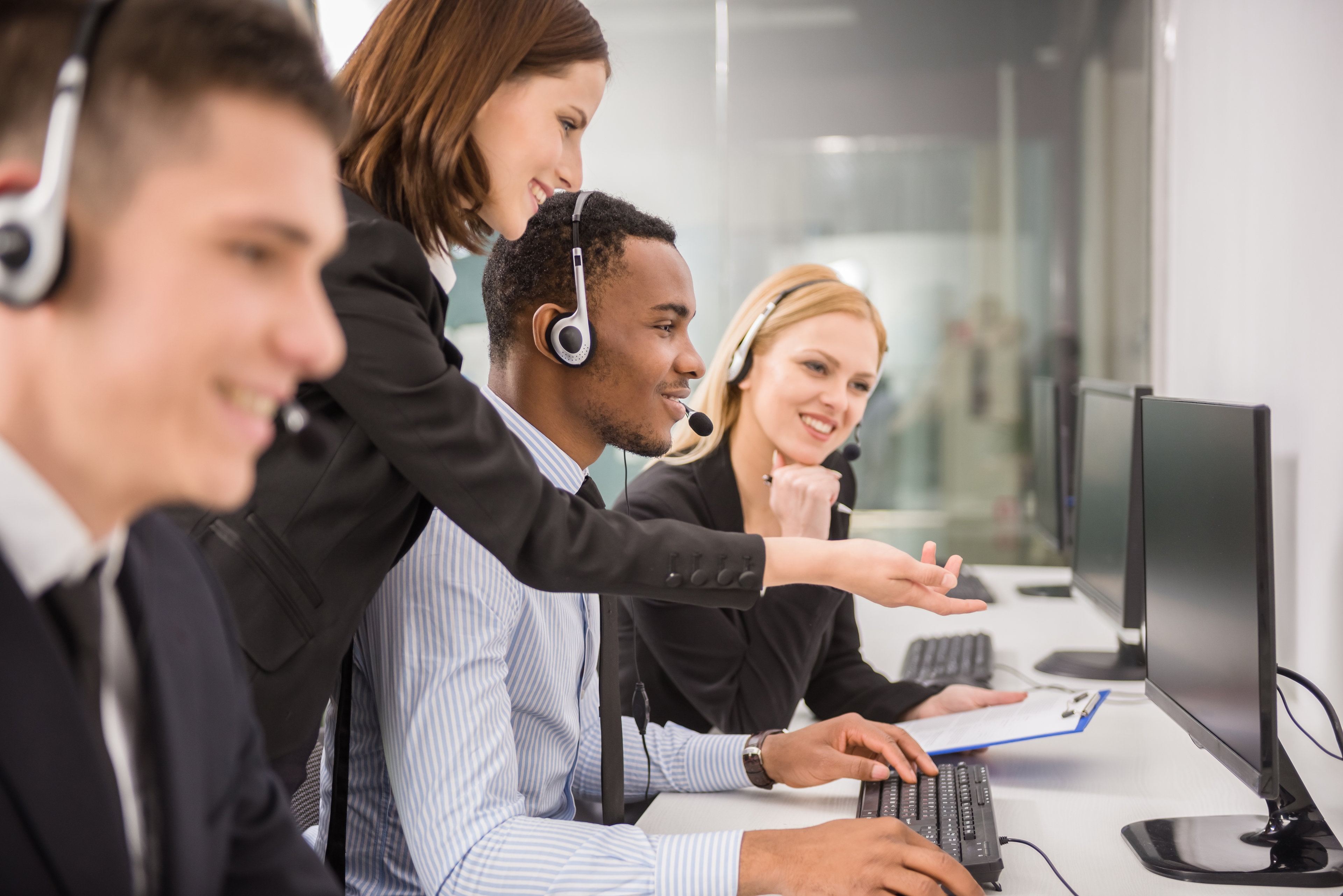 A group of call center workers is getting trained. A woman is gesturing to a computer while a man looks at said computer with his hands on the keyboard and mouse. Another woman is off to the side with a clipboard.