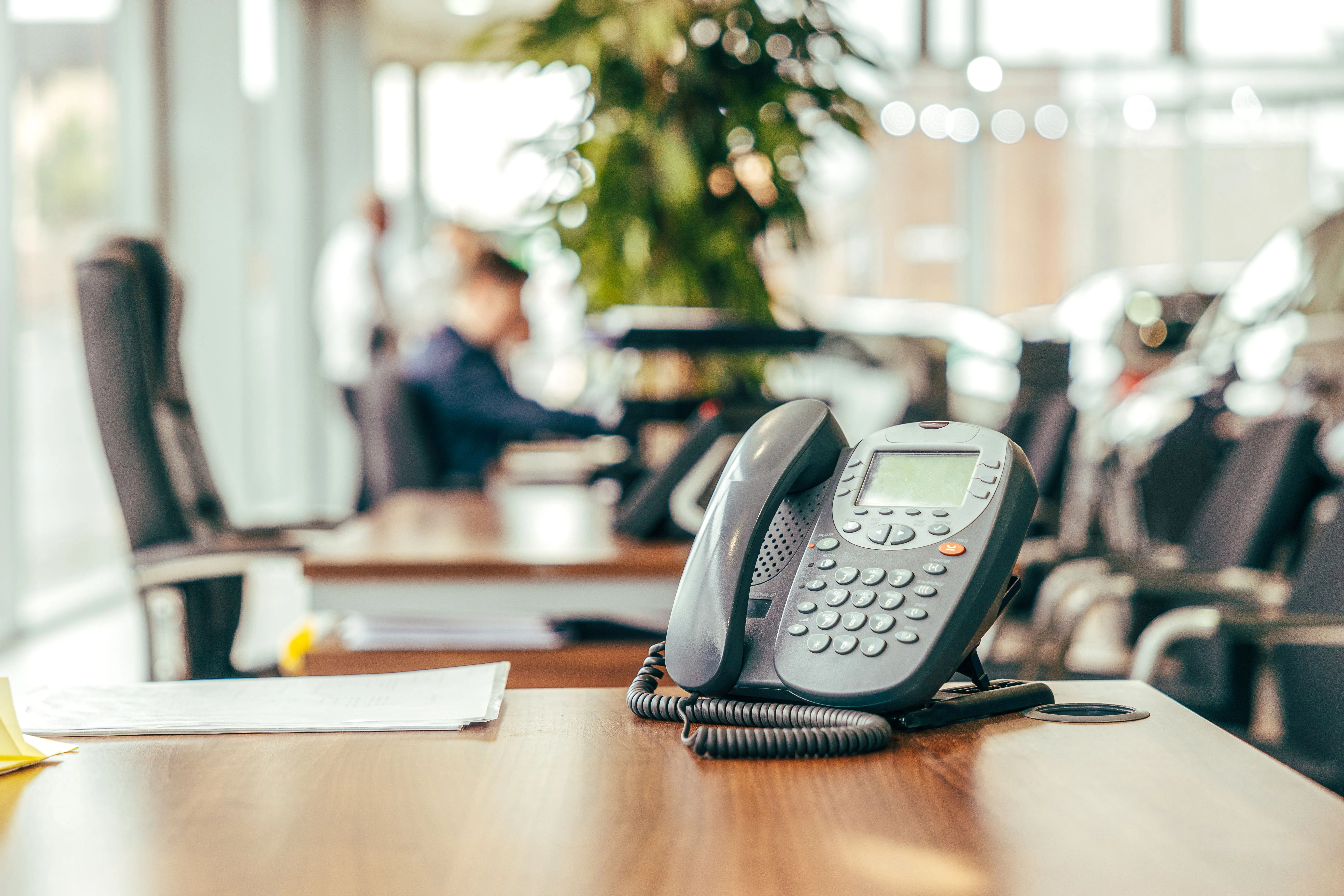 A black office phone sat on a mahogany desk. There are office chairs, plants, and office workers in the background.