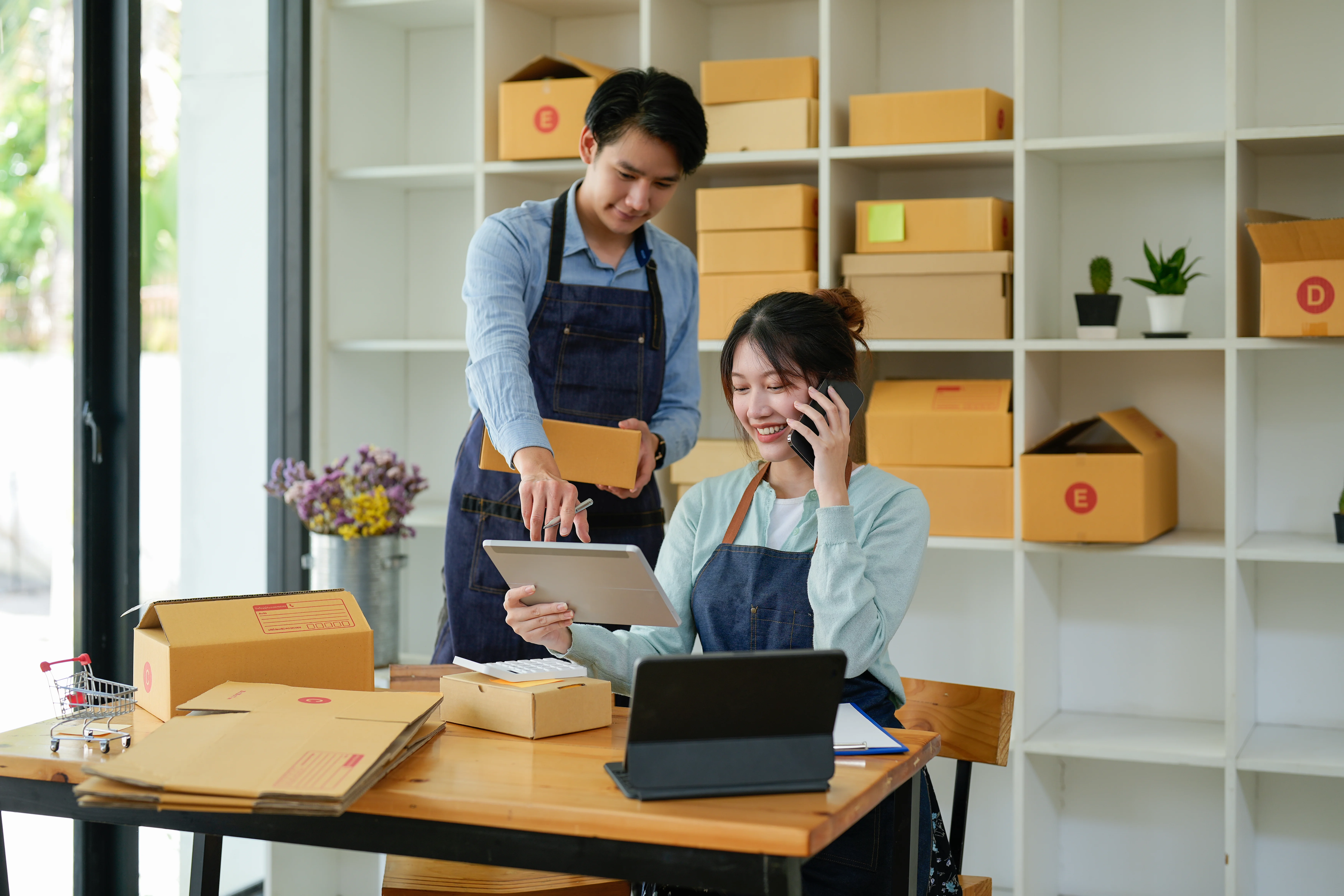A woman and a man work together in a bright, organized workspace. The woman, with her hair in a bun, is on a smartphone call, and using a tablet, while the man stands beside her, holding a small parcel. They're surrounded by cardboard boxes, some marked with letters, and a white shelving unit behind them holds more boxes and a few plants. The scene is well-lit, evoking a light_hearted and efficient atmosphere. This image could be used for advertising online stores, small business partnerships, shipping and logistics, or technology solutions for entrepreneurs.