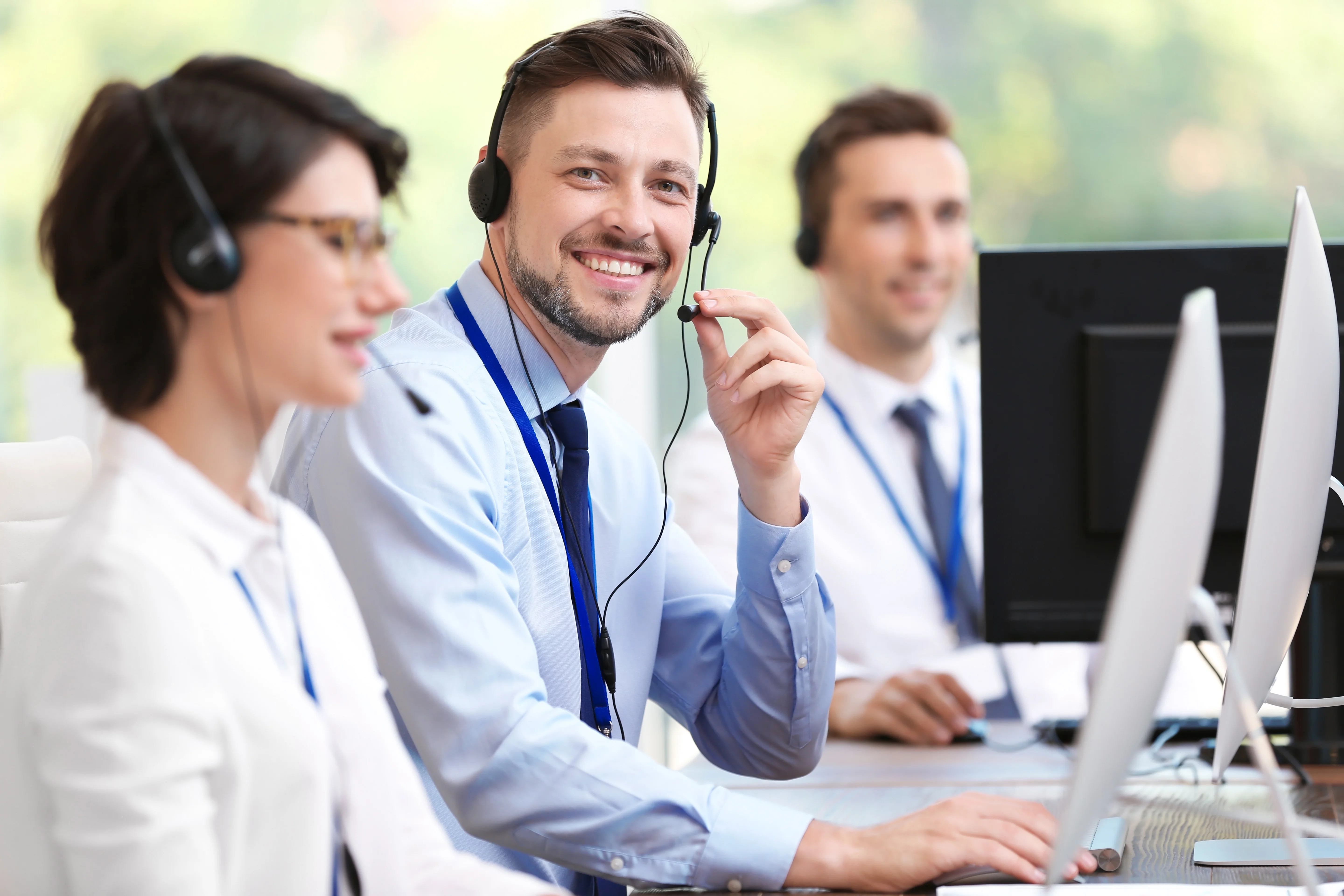 Three friendly customer service agents are shown in a bright, modern office setting. Each subject is wearing a headset, indicating they are ready to assist customers. Two men and one woman are at their desks, each with computer monitors in front of them. One man in the foreground smiles directly at the camera while holding his headset's microphone. The office has a bright, clean appearance. This image is perfect for portraying customer service, support, professionalism, and communication.