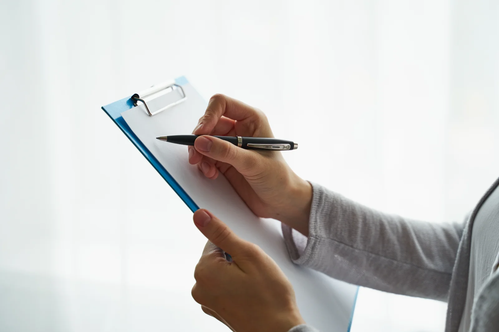 Close-up of a hand holding a pen and writing on a blue clipboard with white paper against a light, out-of-focus background. The individual is wearing a gray top. This image evokes professionalism and focus, and could be used for projects related to business, healthcare, or education. The clean, simple composition lends itself to use as a background or overlay in documents and presentations.