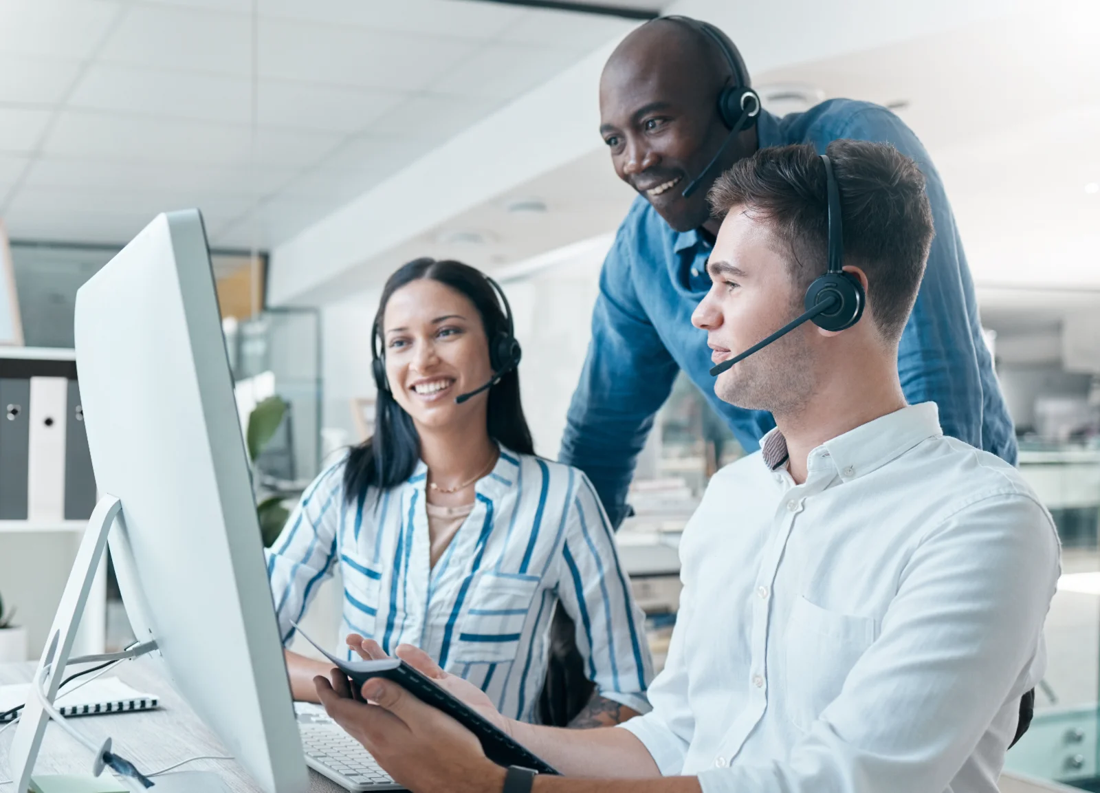 Three colleagues wearing headsets are working together at a computer in an office. The woman with long, dark hair is smiling as she looks at the screen. One man is seated and wearing a white collared shirt and headset. Another man with short dark hair is wearing a blue shirt and headset. They are working at a desk in a brightly lit office. The scene conveys a lighthearted and collaborative environment. This photo would be useful for projects needing to portray teamwork, customer service, or modern workplace settings.