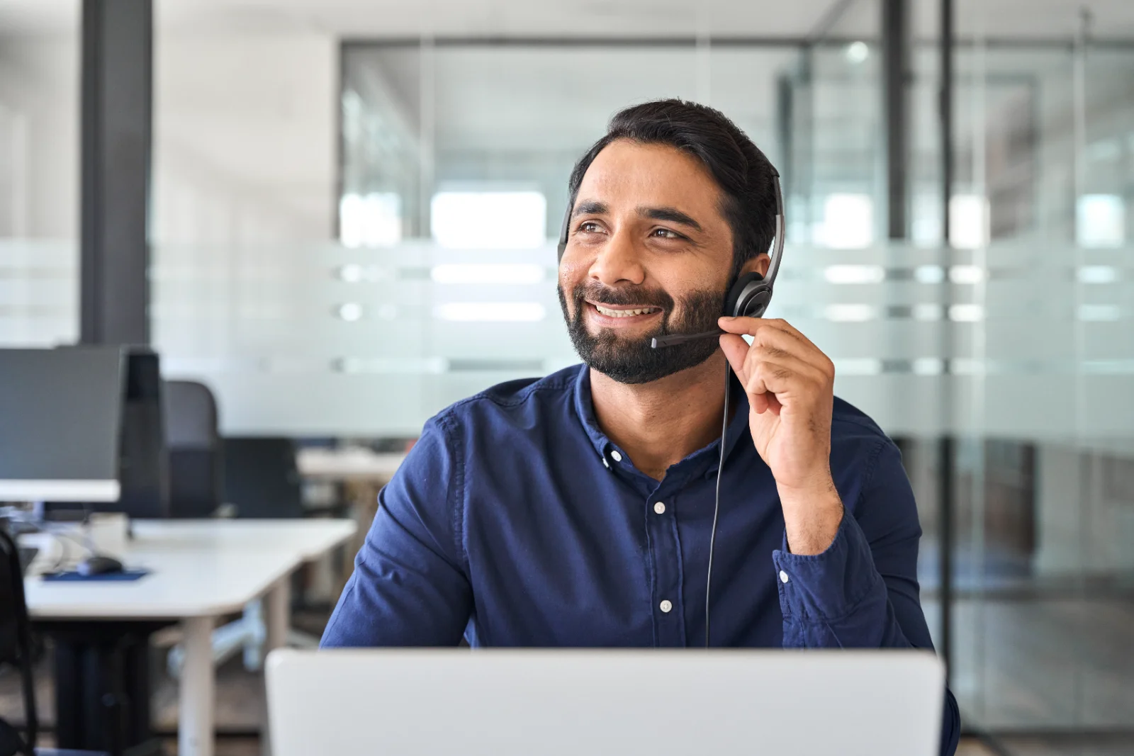 A man with short dark hair and a beard sits at a desk wearing a headset in a modern office. He is smiling and looking to the side as if talking to someone. The man is wearing a blue collared shirt. In front of him is a white laptop computer. The background shows a bright office with desks and glass walls. This image could be used for projects about customer service, technology, or the workplace.