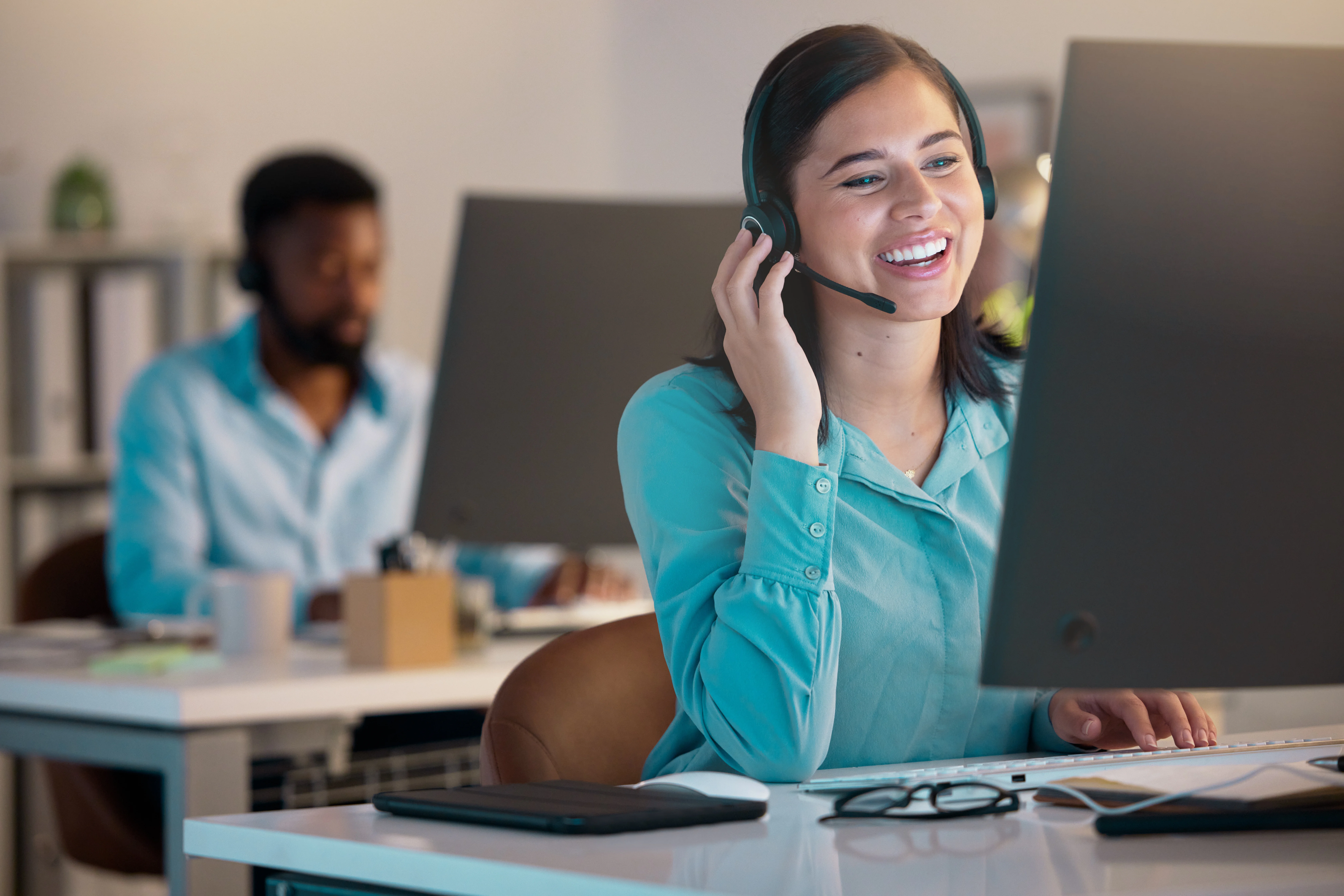 A woman using a headset to talk to someone. She is smiling at her computer and there is a man in the background.