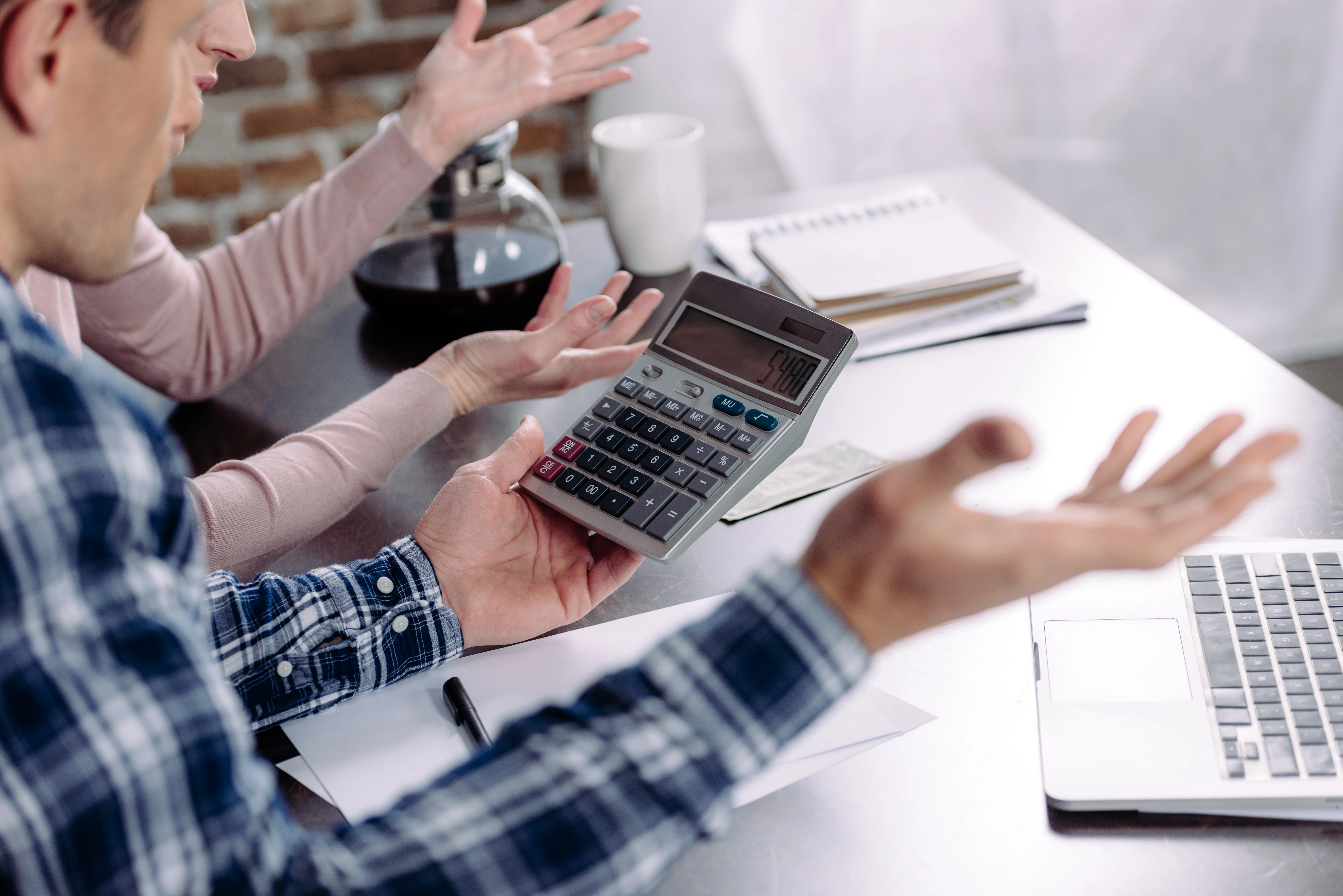 A frustrated couple adding numbers. They have a calculator and a pot of coffee in front of them.