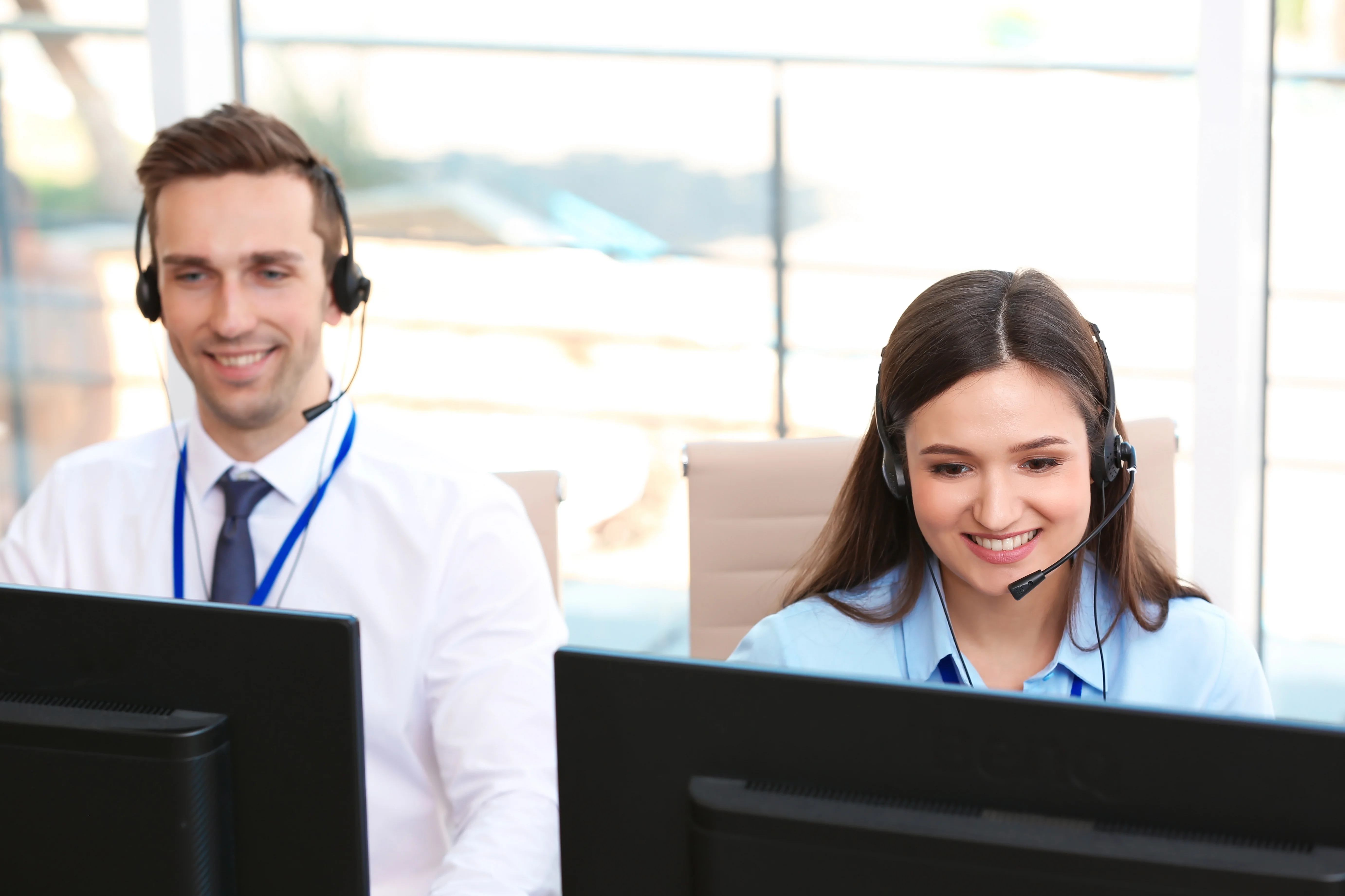 Two coworkers at a call center sitting next to each other. They are wearing headsets and looking at computers.