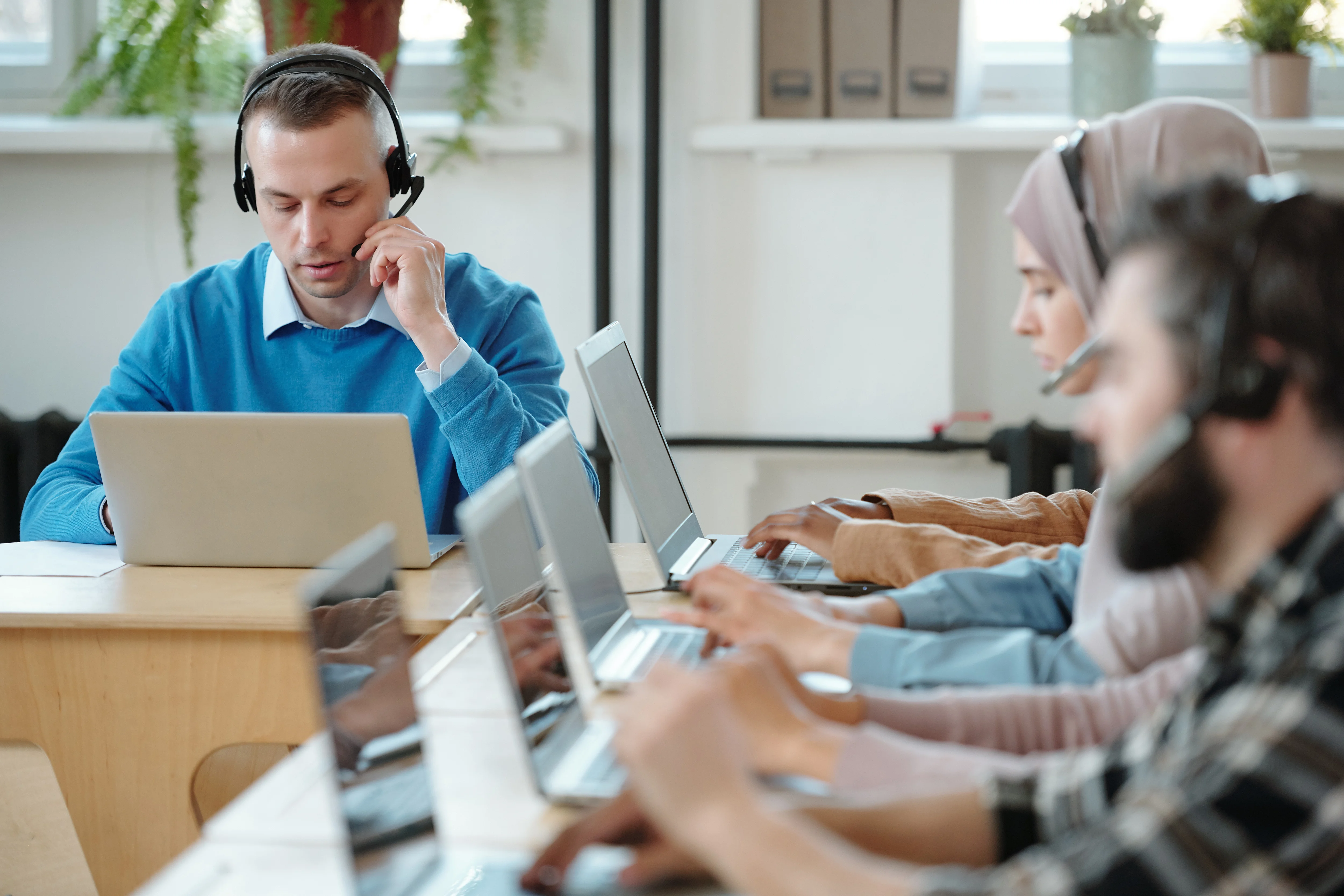 Four adults are working on laptops at desks in a modern, bright office. The office has large windows and is decorated with plants. The people are wearing professional clothing. The scene shows a productive and collaborative environment. This photo can be used for projects about customer service, call centers, business, technology, and modern work environments.