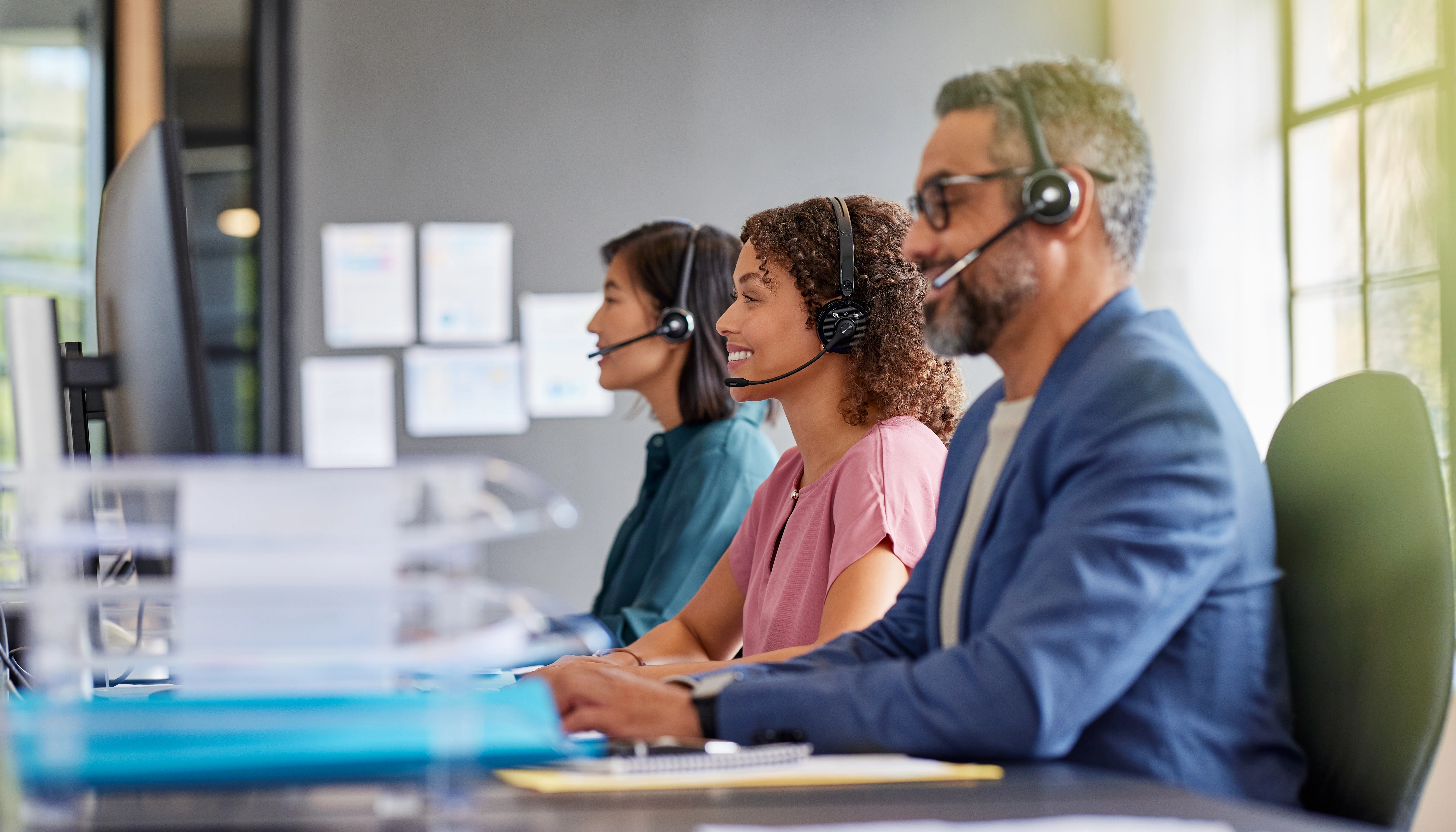 Three people sit at desks in a modern office, wearing headsets and working at computers. The office has a neutral color scheme and natural light from the window. They appear focused and professional, creating a sophisticated and efficient working environment. This image is perfect for use in corporate communications, advertising, or websites related to customer service, technology, or the modern workplace.