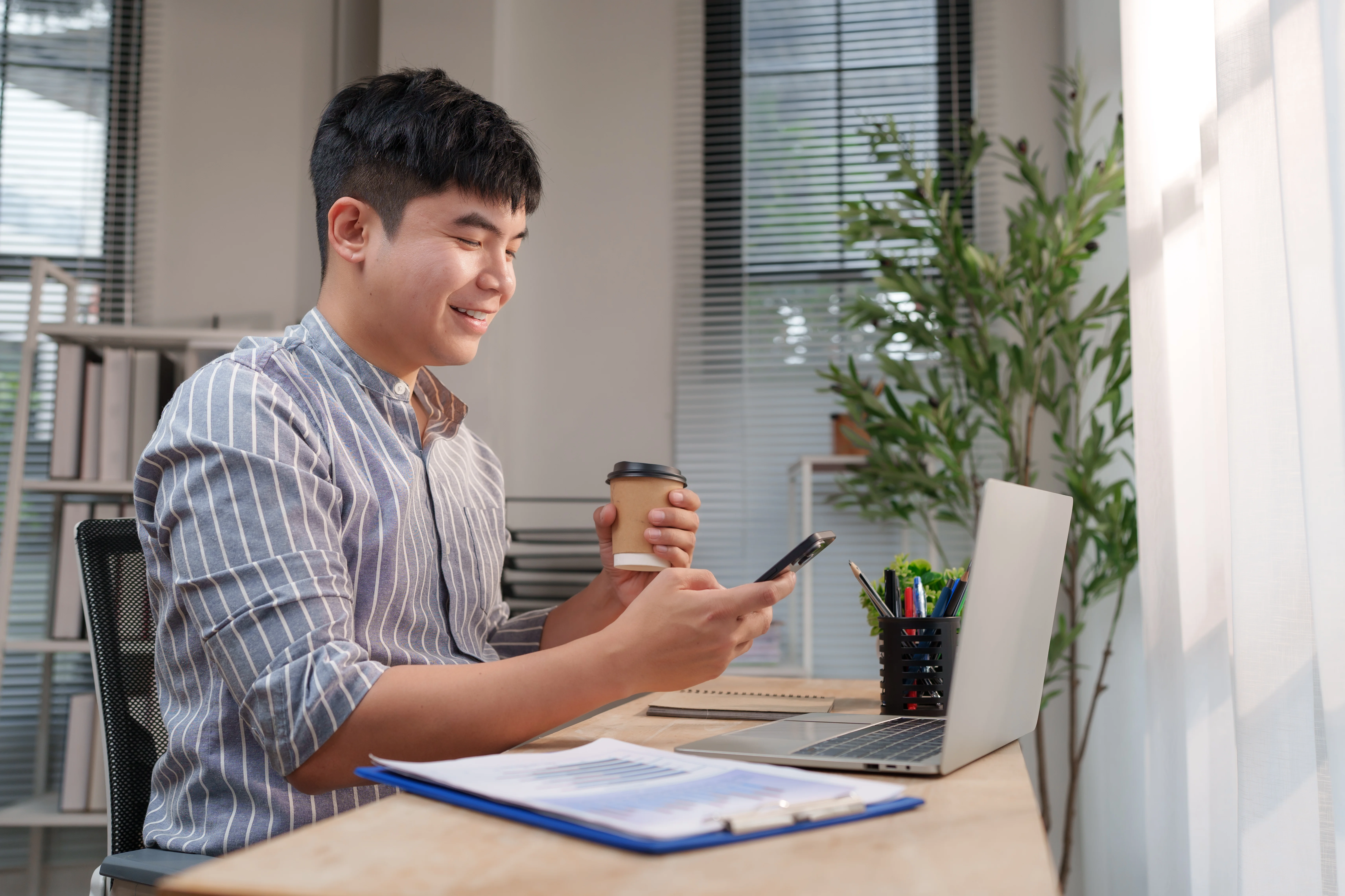 A smiling young man sits at a light-colored wooden desk in a bright, modern office or home workspace. He is wearing a blue and white striped button-up shirt and has short black hair. In one hand, he holds a disposable coffee cup, while the other hand operates a smartphone. On the desk, a laptop sits open next to a container of pens and some documents on a blue clipboard. The room is filled with soft, natural light coming through a window with white curtains and blinds, creating a productive atmosphere. This authentic image is ideal for projects centered on remote work, modern business culture, technology, productivity, and balanced lifestyle.