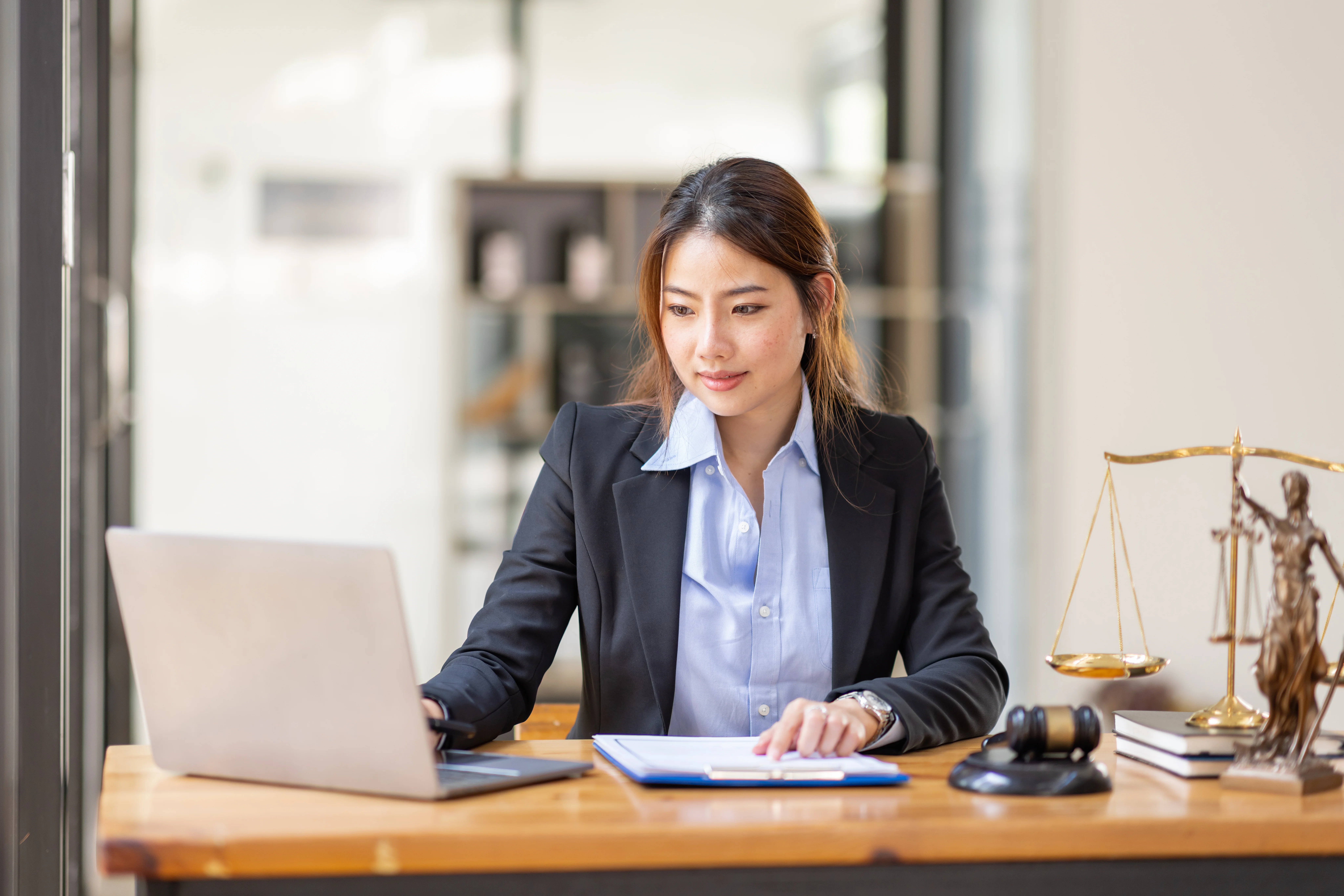 A woman looking at her computer with a clipboard in front of her. She has a gavel and scales next to her.