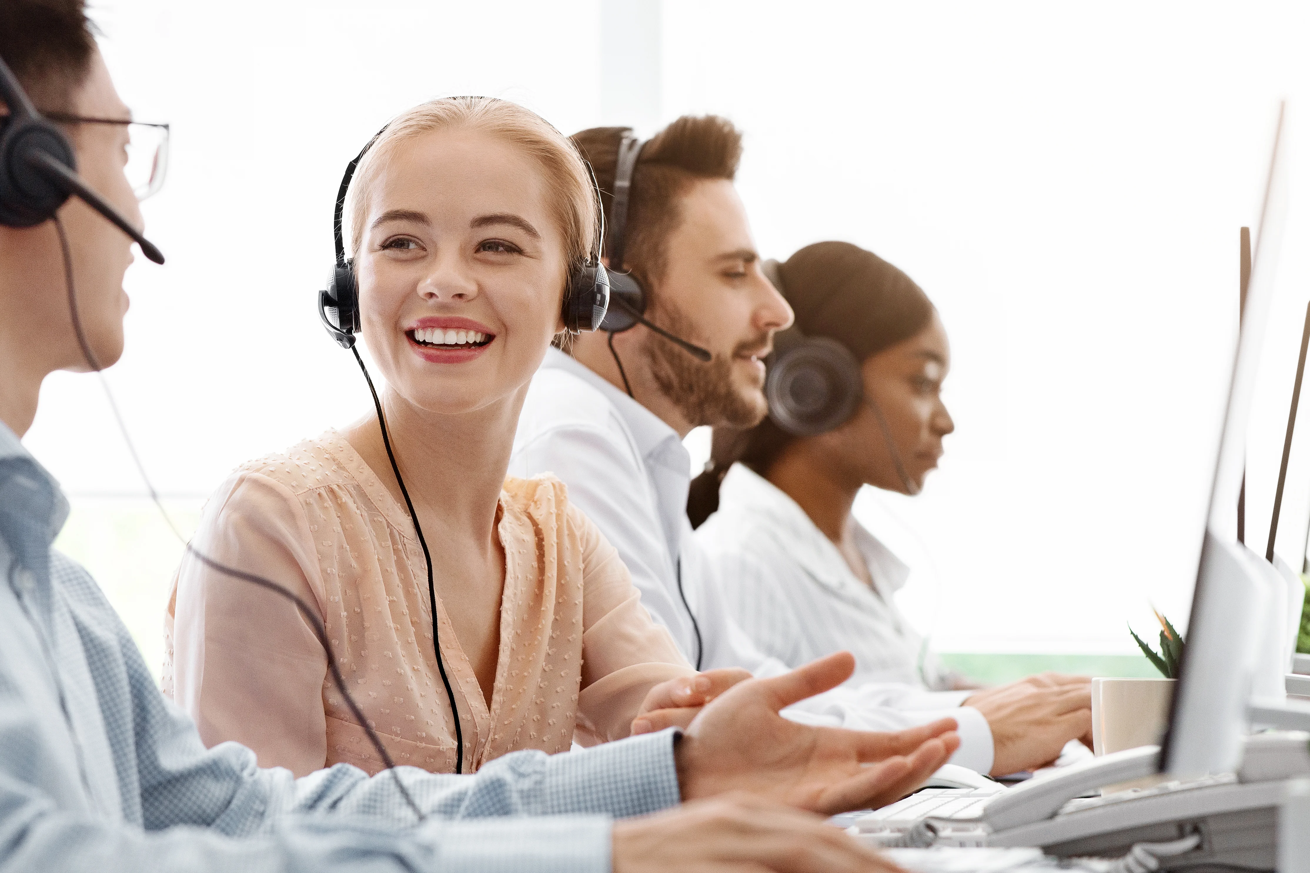 A woman and her coworker talking. Both are wearing headsets, and they are sitting in front of their respective computers. There are two people in the background who look like they are having conversations with customers.