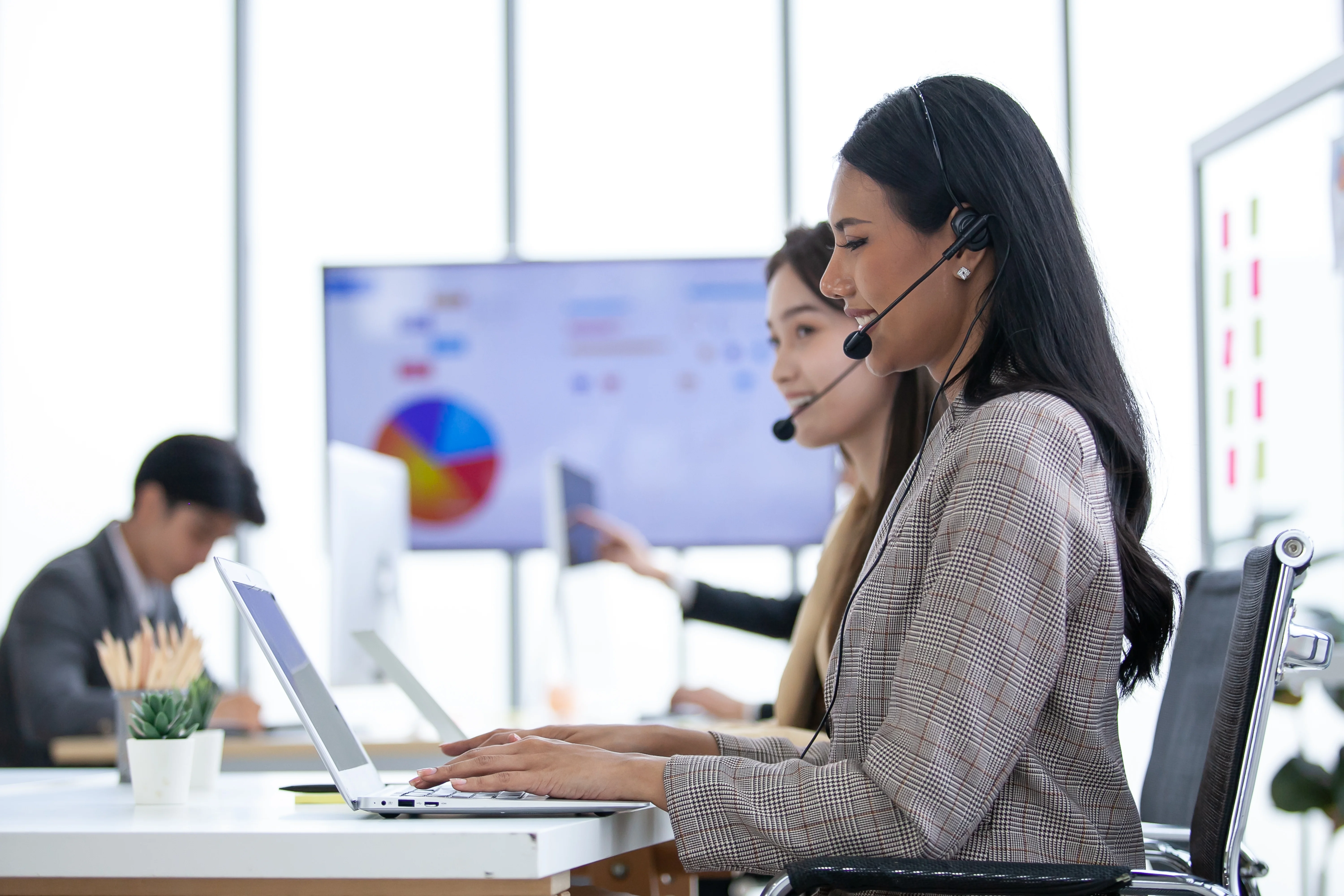 A group of coworkers sitting at a collaborative desk. Two women are wearing headsets and talking to customers through their cloud phone system. There are several graphs and charts on a screen behind them.