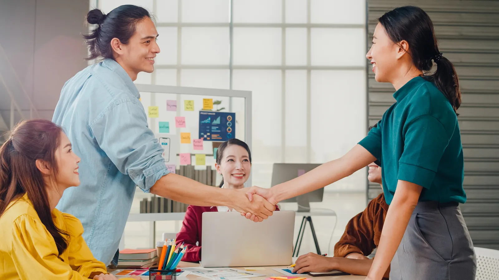 A man and woman shaking hands across a table. They seem to be in a business meeting with other folks around the table with them.