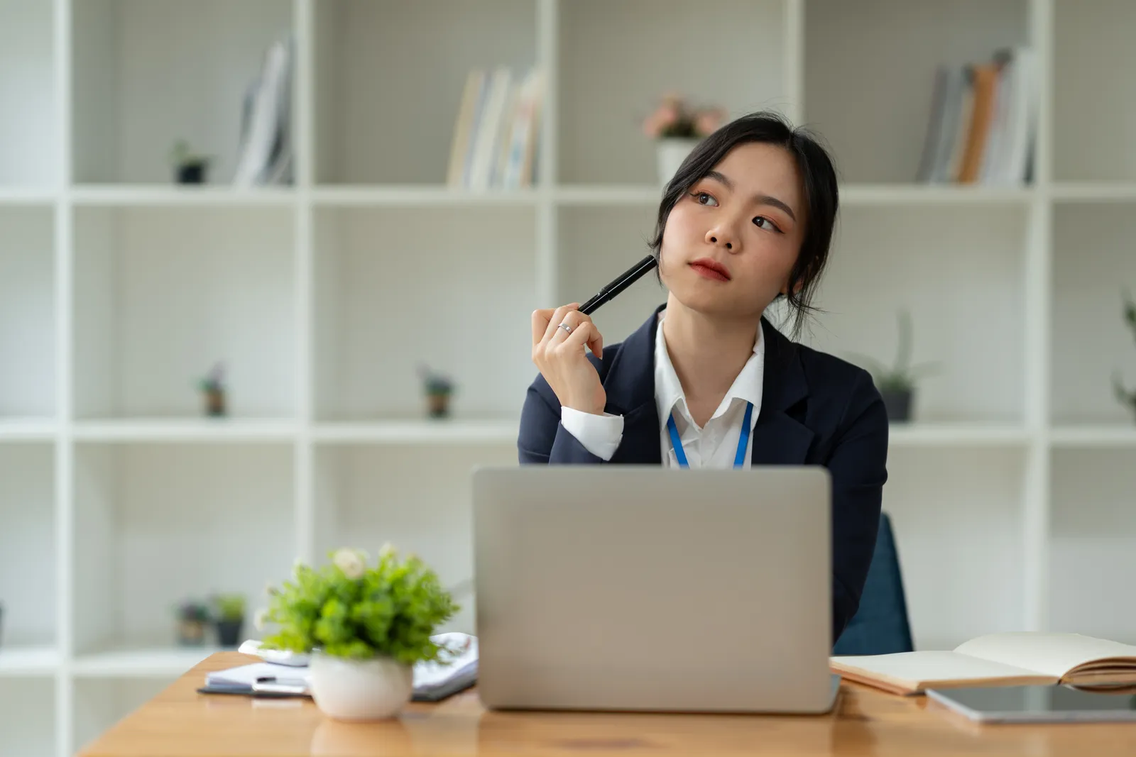 A woman sitting at a table with a laptop, clipboard, and notebook in front of her. She has her pen pointed towards her face as if she is in deep thought.