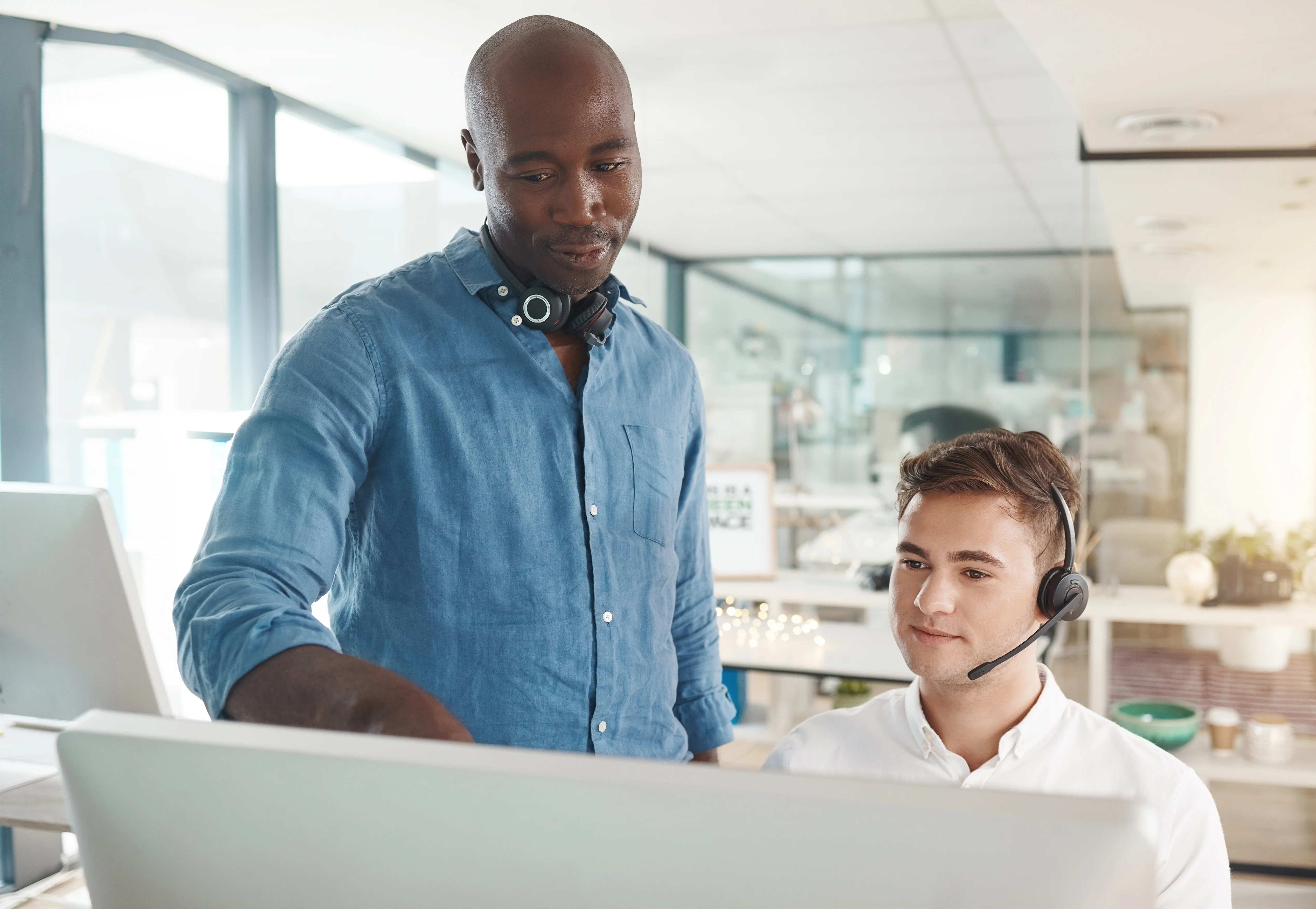 Two men working in an office together. One of them is training the other. He is pointing at the computer while his colleague is wearing a headset and staring at the computer screen.