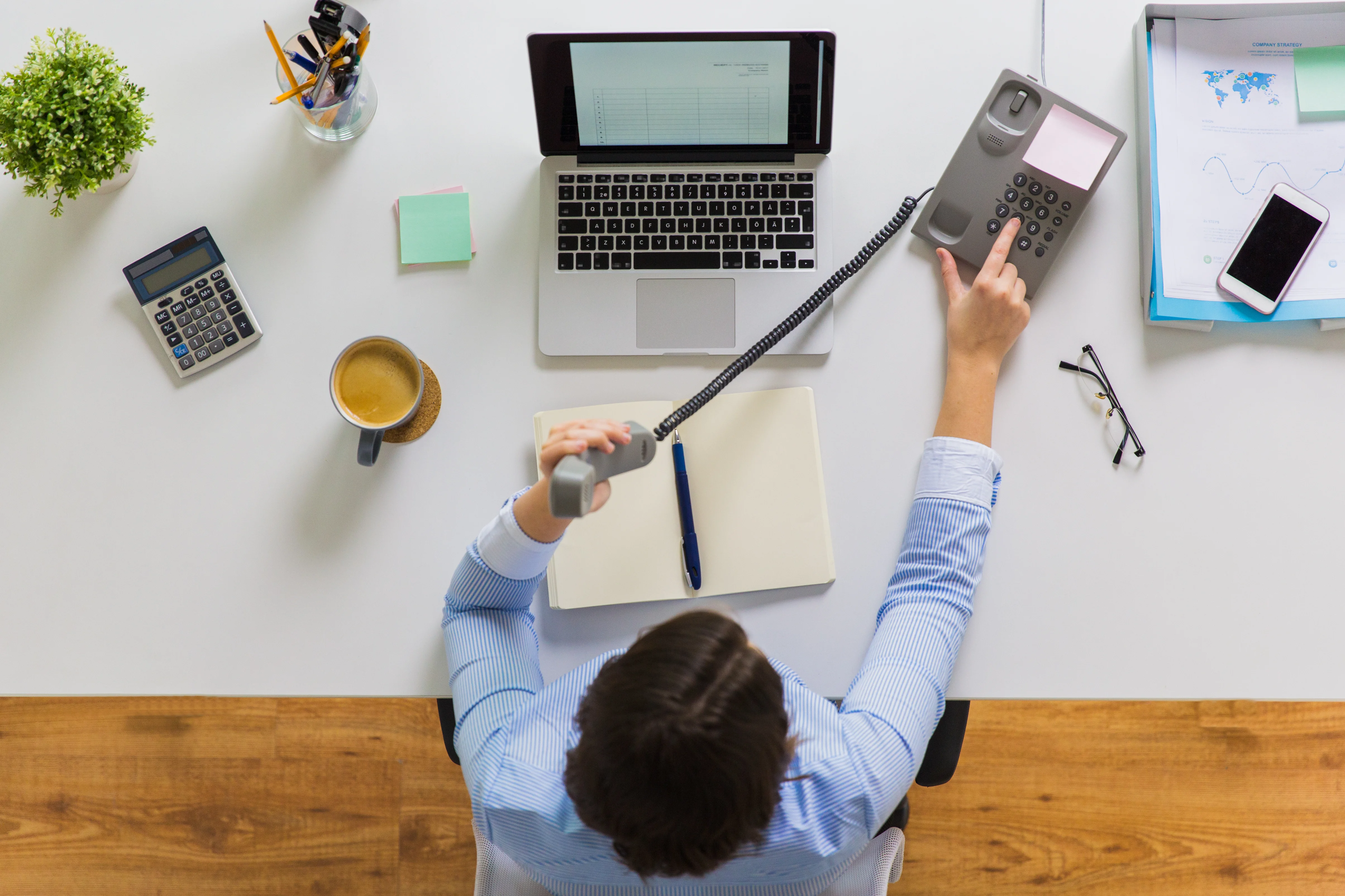 A woman making a phone call on a traditional phone system. She has a notebook and laptop open on her desk. There is a cup of coffee, sticky notes, a calculator, pencil cup, glasses, and a phone atop her desk.