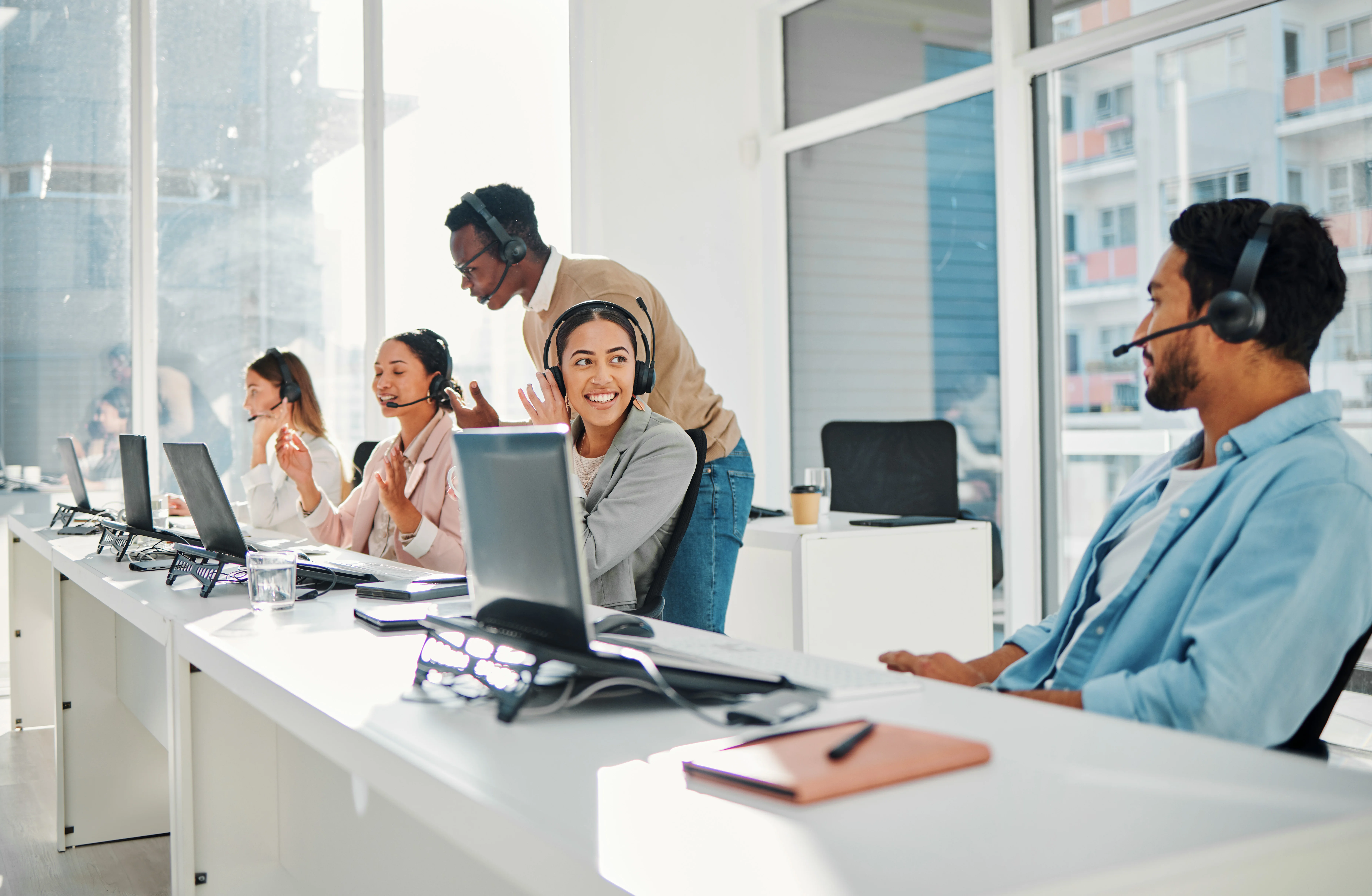 A group of coworkers wearing headsets. Two of them are talking to each other and another is getting trained by a manager. One person in the background is on a call.