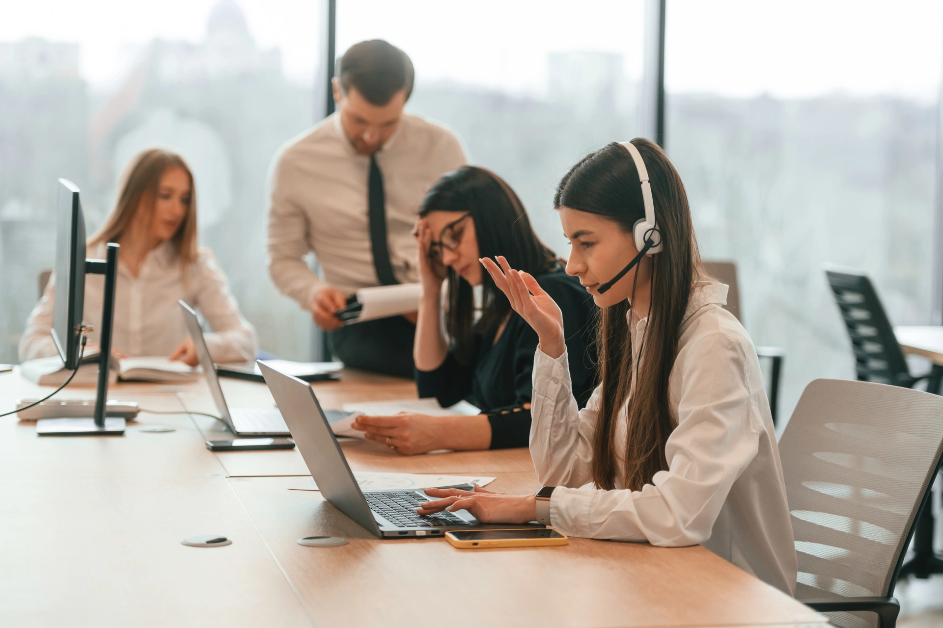 A group of coworkers working in a collaborative space together. Three women are sat down on chairs while a man is leaning against the table. Two women in the foreground are visably frustrated with both seemingly exasperated at the situation.
