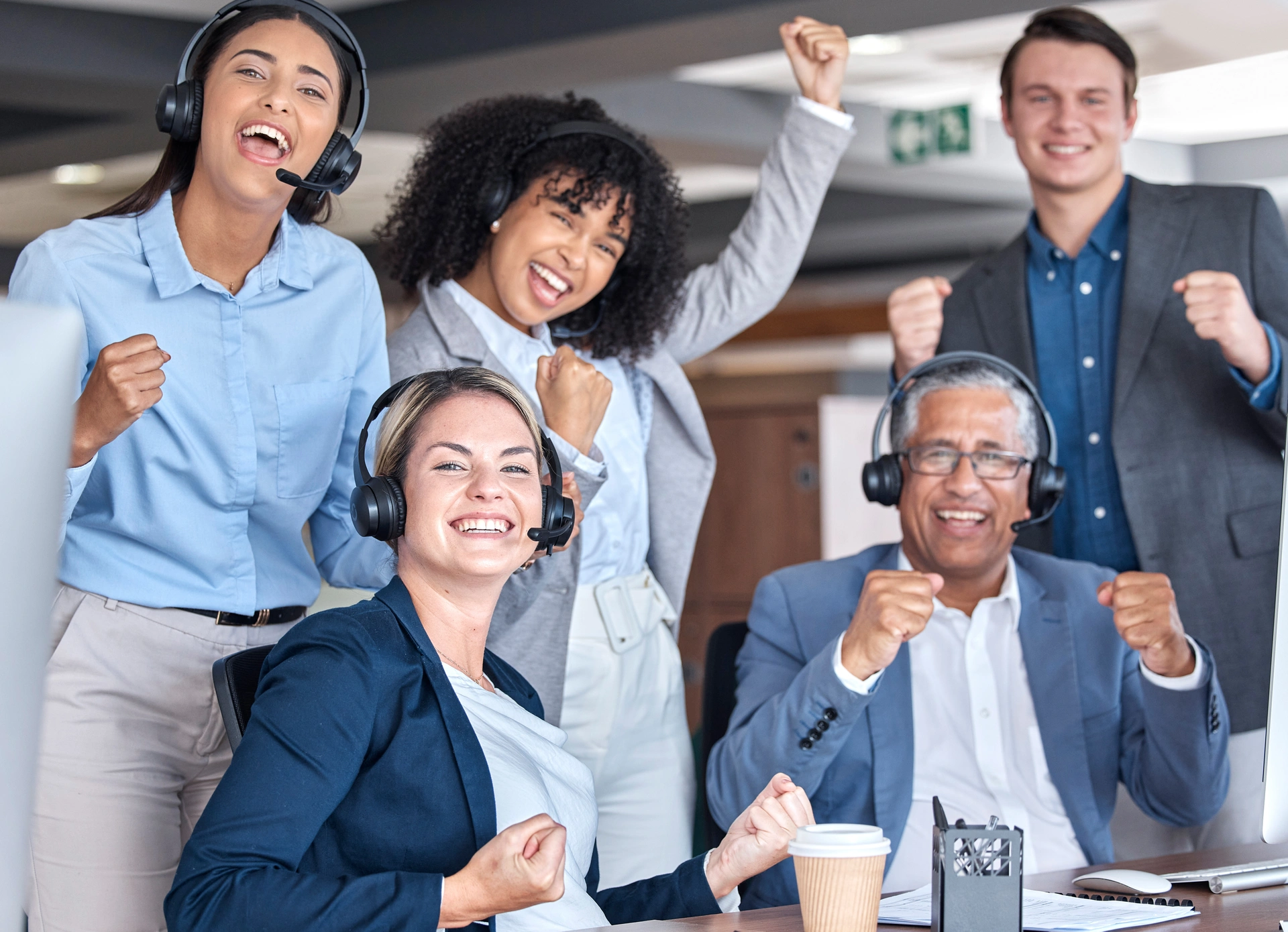A group of coworkers cheering and looking directly into the camera. Four of them are wearing headsets, and one man in the background isn't. They are all enthusiastically looking into the camera.
