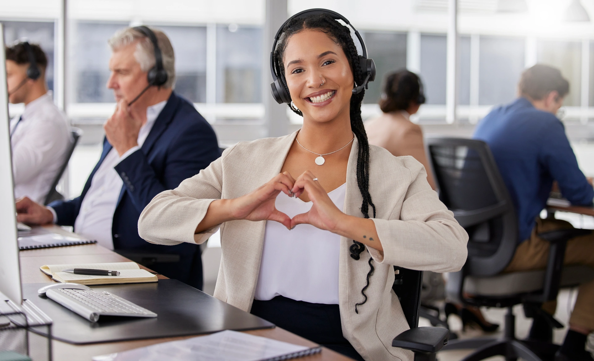 A woman at a call center. She is looking into the camera with her headset on. She has her hands formed in the shape of a heart.