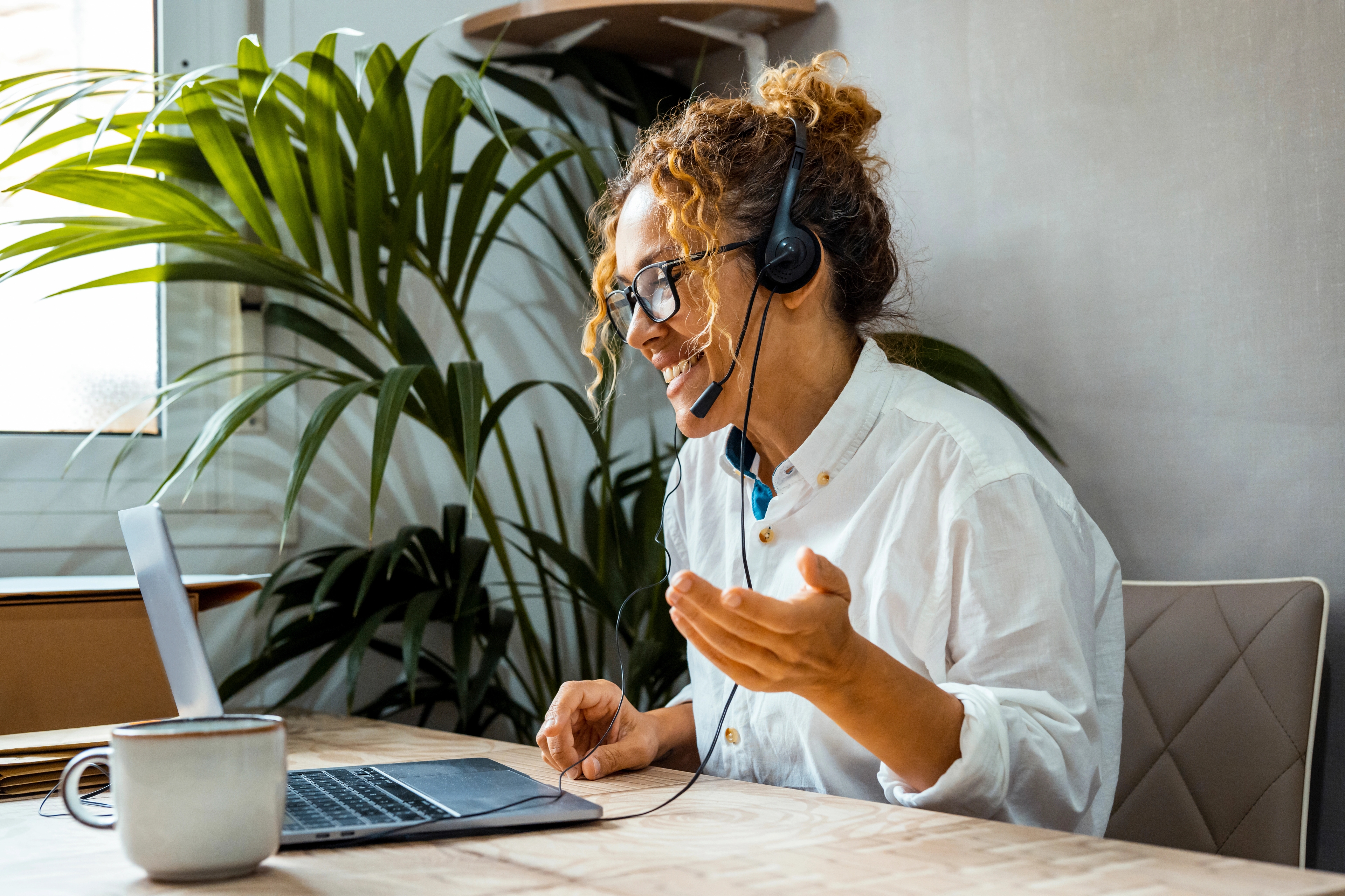 A woman at home on a headset that is connected to the laptop in front of her. She is enthusiastically talking.