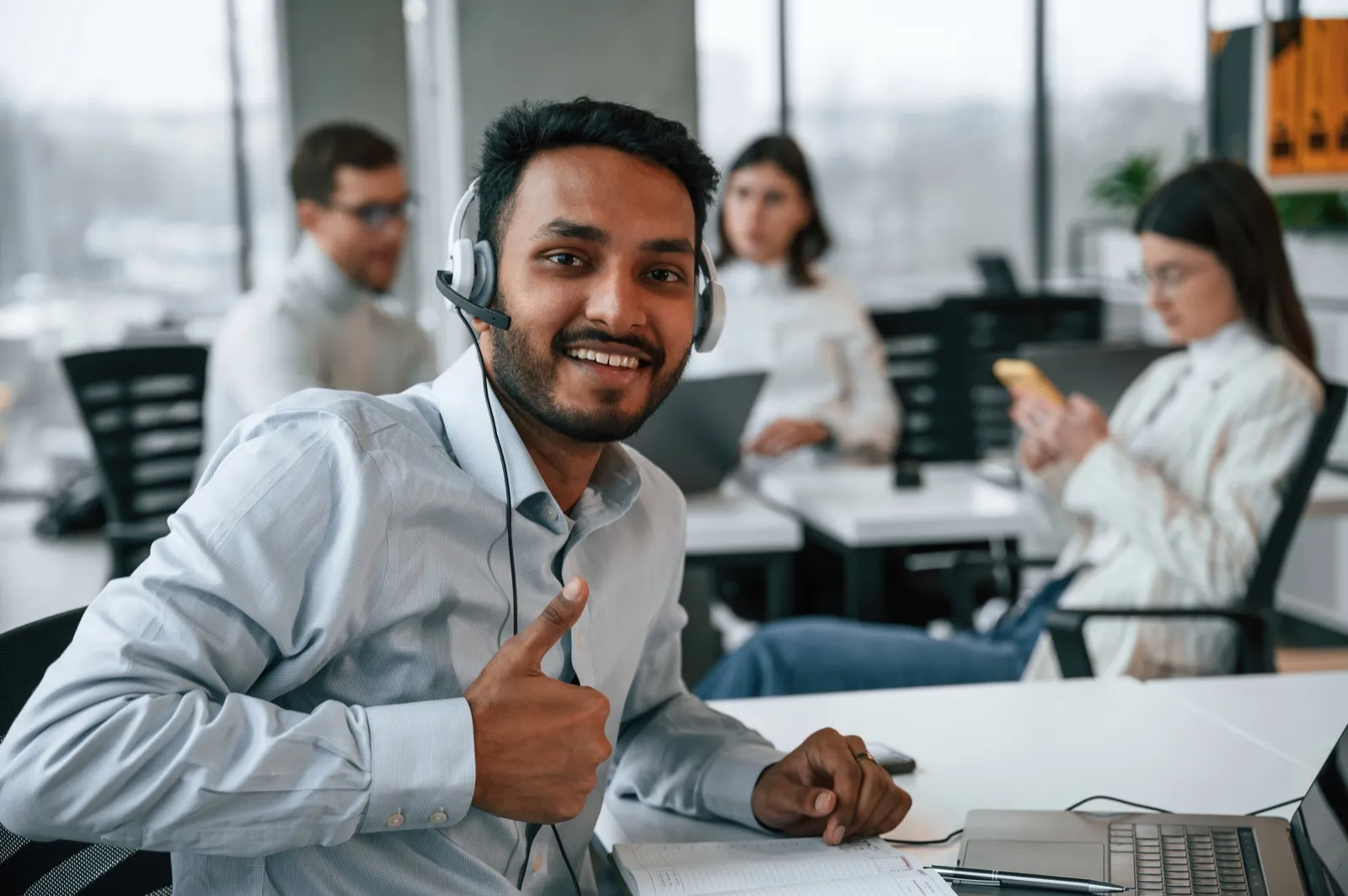A man wearing a headset looking into the camera and smiling. There are three coworkers blurred in the background.