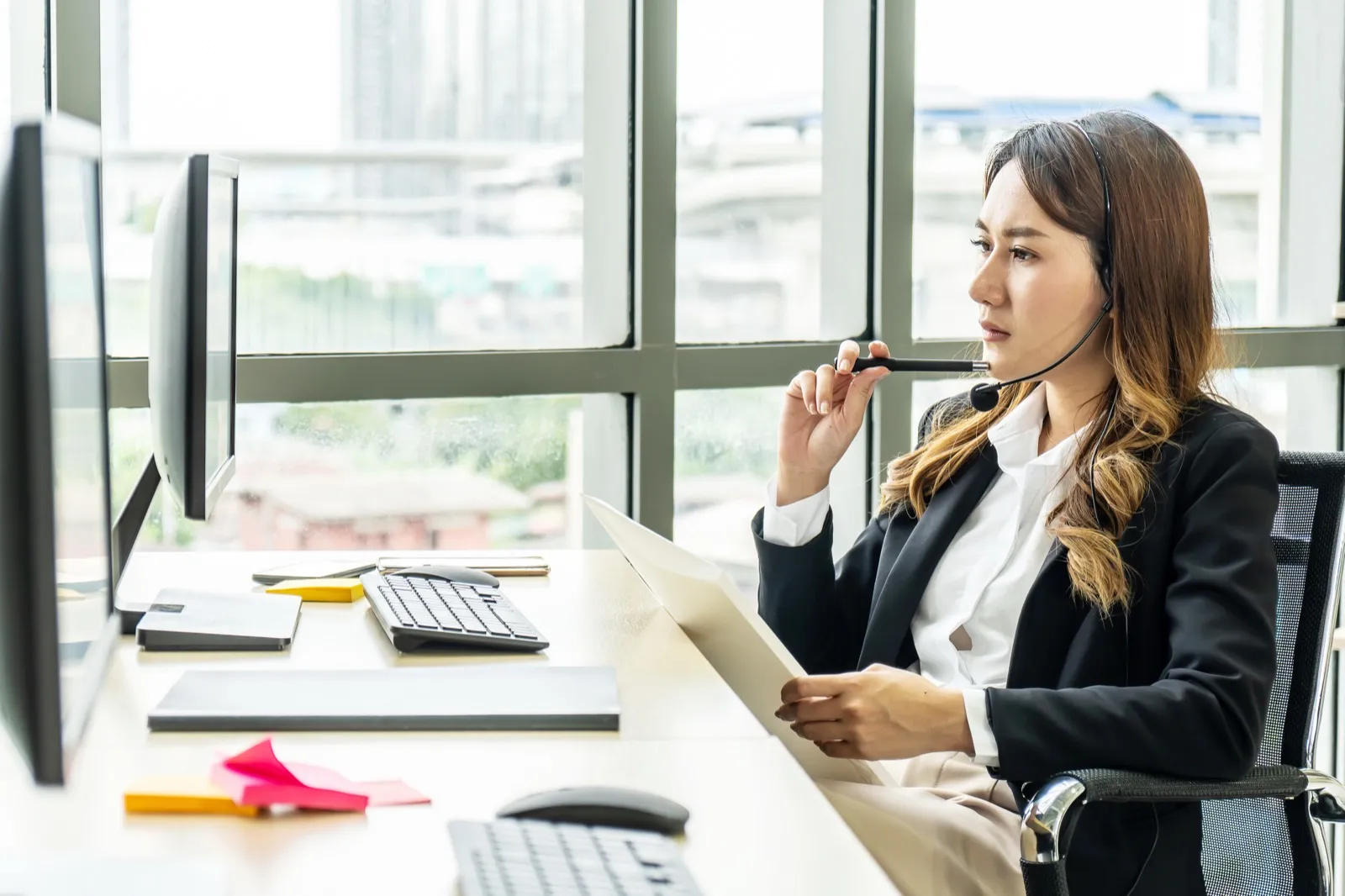 A woman looking at the computer. She is wearing a headset and has a pen to her chin. In her other hand, she has a piece of paper.