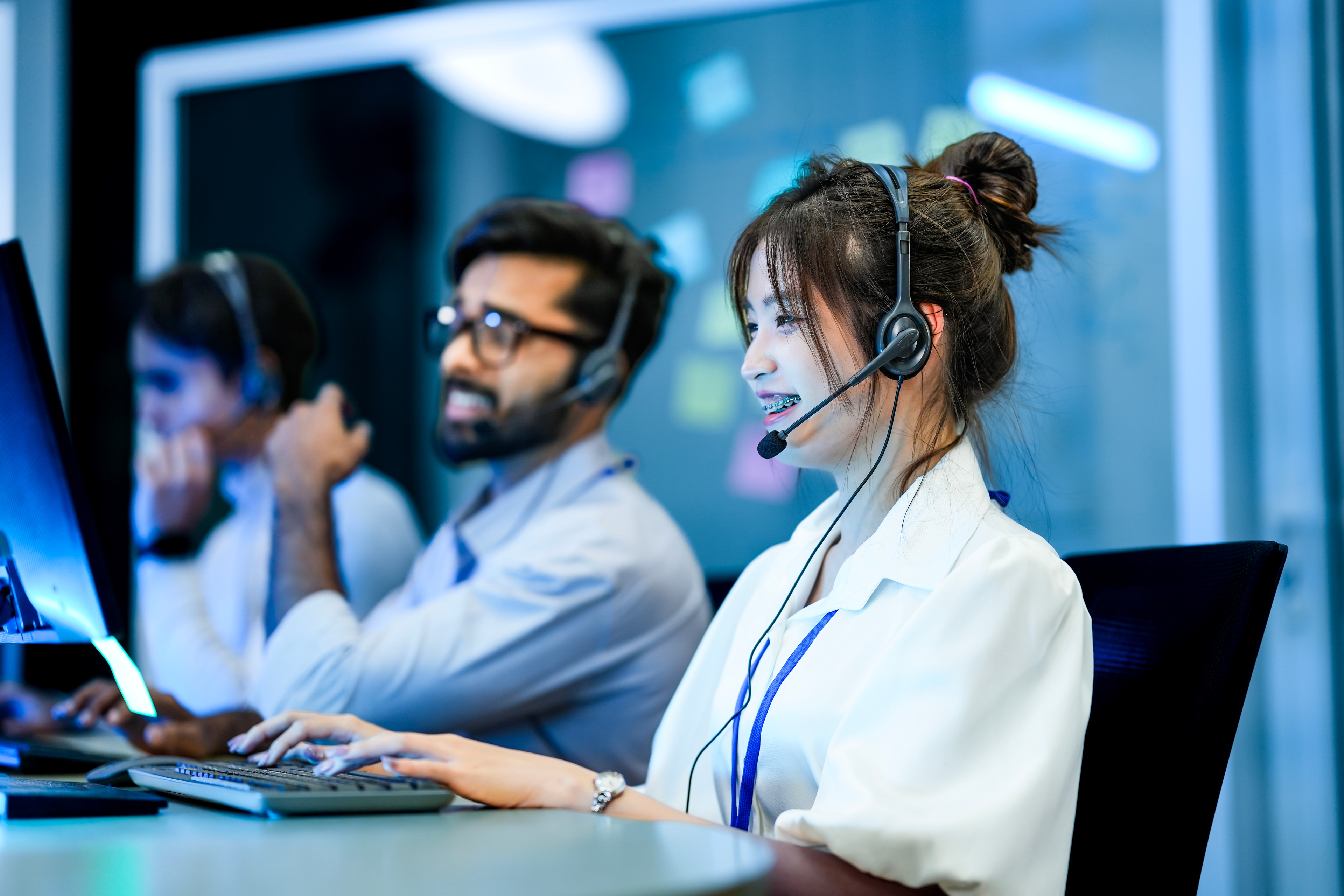 A woman with a headset on. She has her hands on a keyboard and is smiling at her monitor. There are a few other call representatives in the background, blurred out slightly.