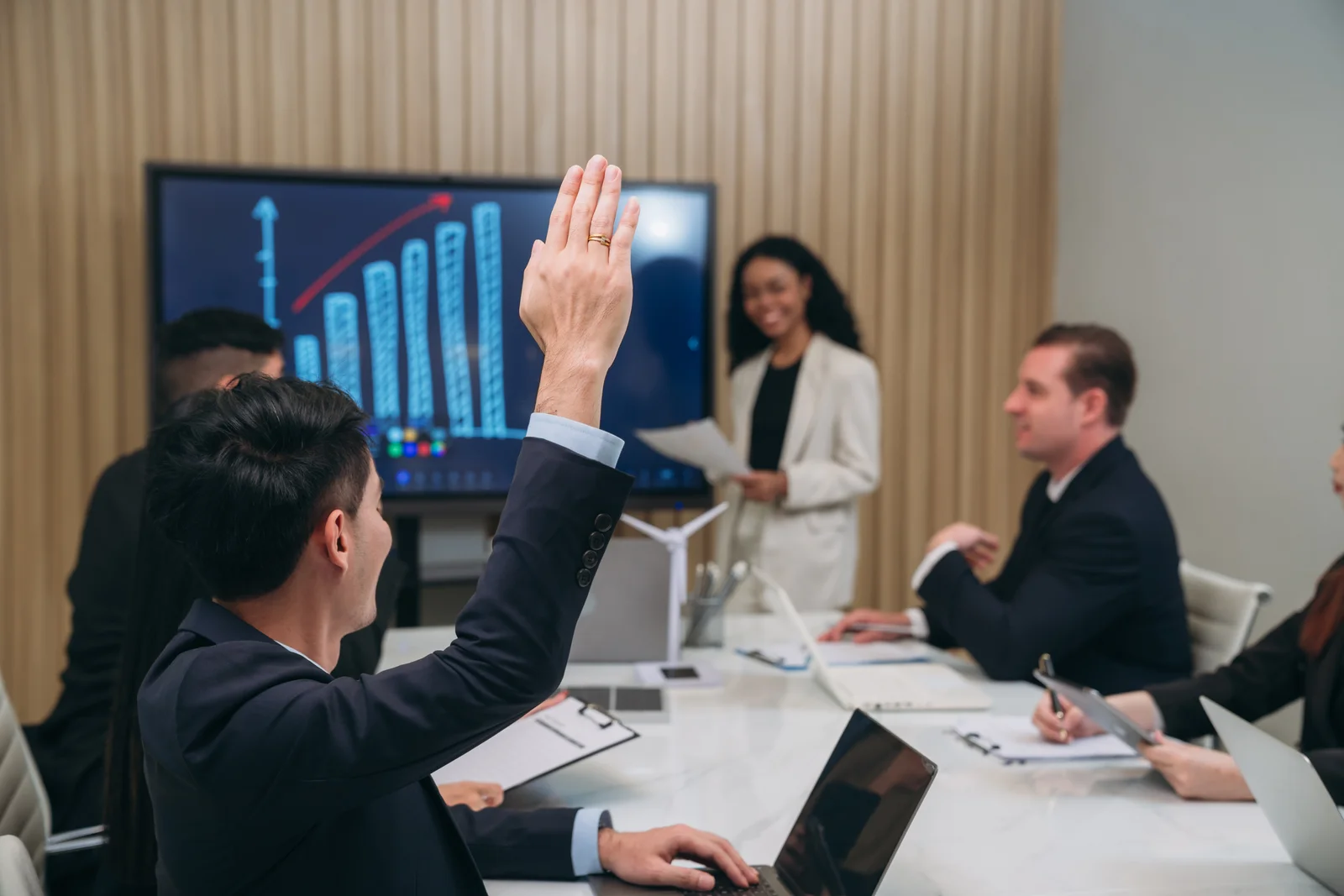A business meeting. The main the foreground has his hand raised and there is graph projected on the screen.