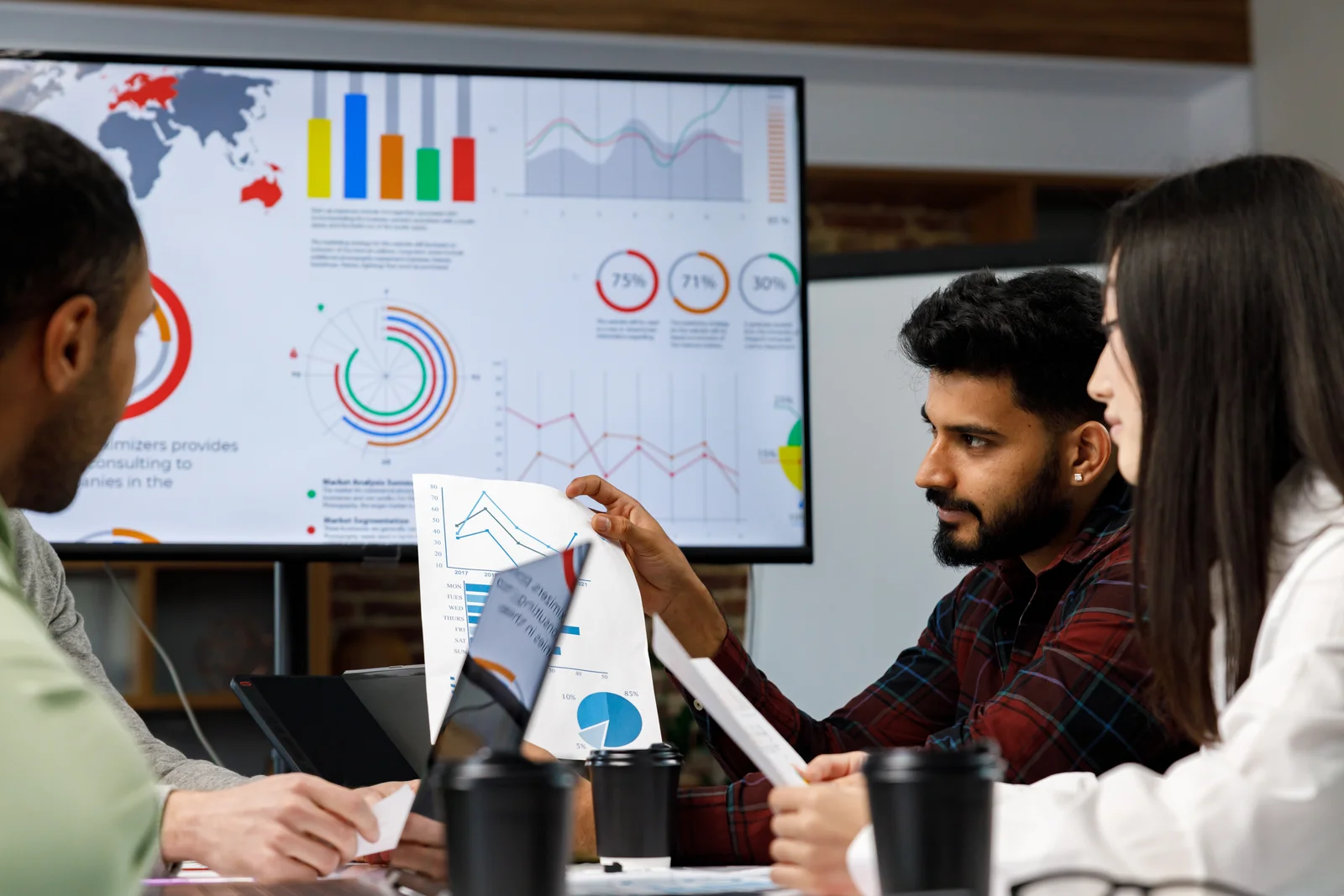 A group of coworkers discussing graphs. One has a graph in his hand and there are graphs projected onto the television screen in the background.