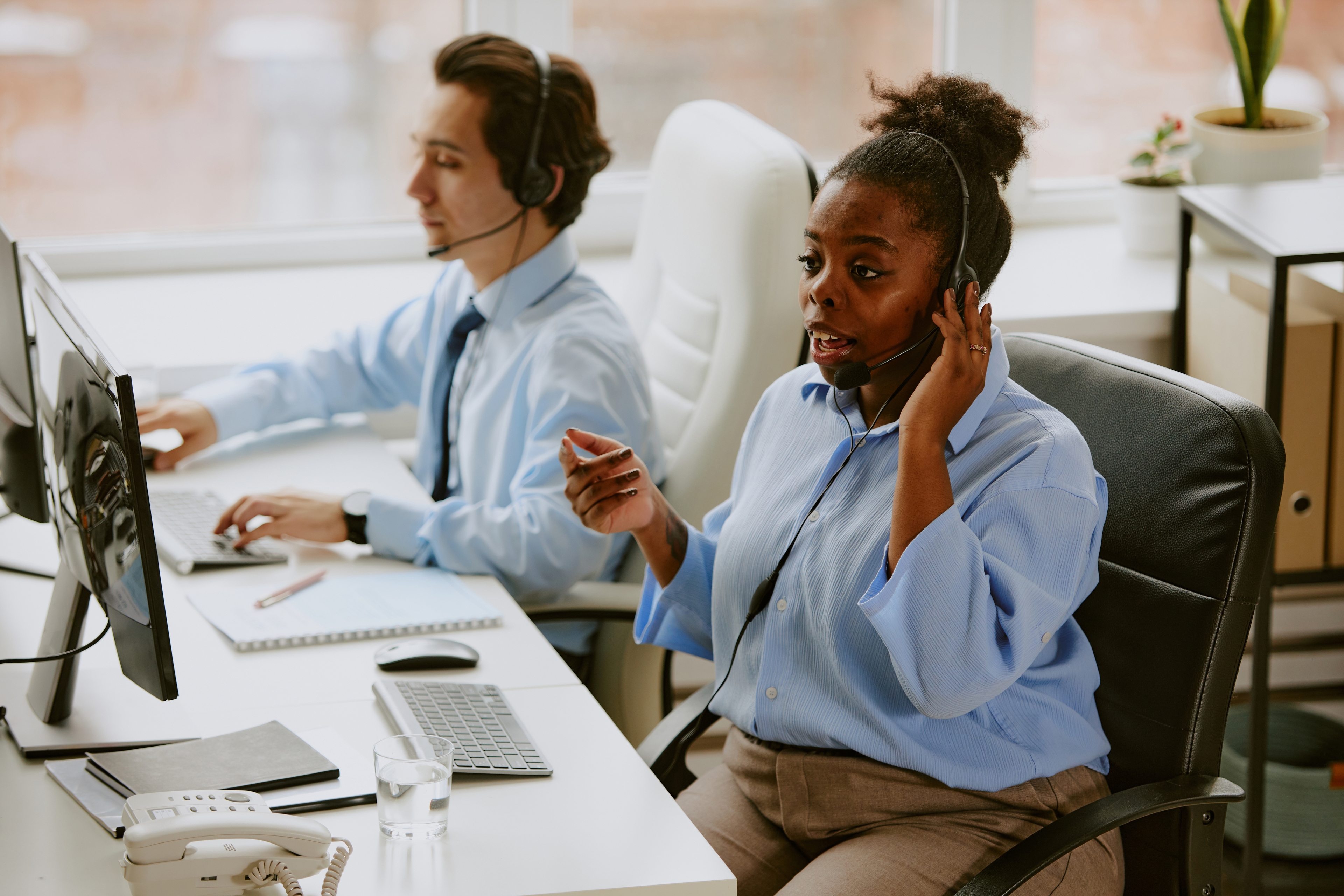 Coworkers talking on their headsets. The woman in the photo is a foot away from the desk talking into her headset explaining something. The man in the background is diligently looking at something on the computer screen.
