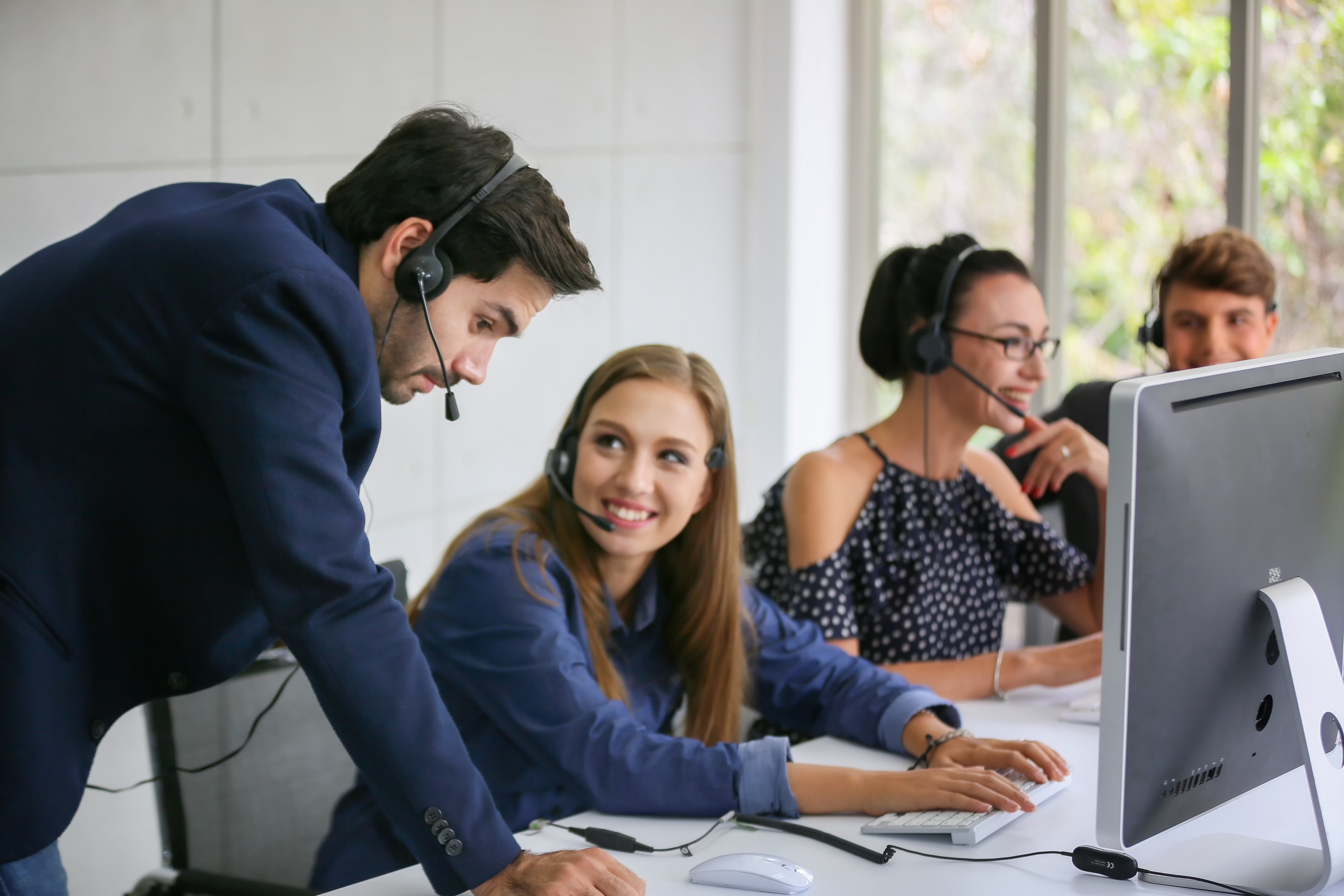 A group of call agents sitting at a communal desk. One woman is getting trained by a man who is leaning on the desk.
