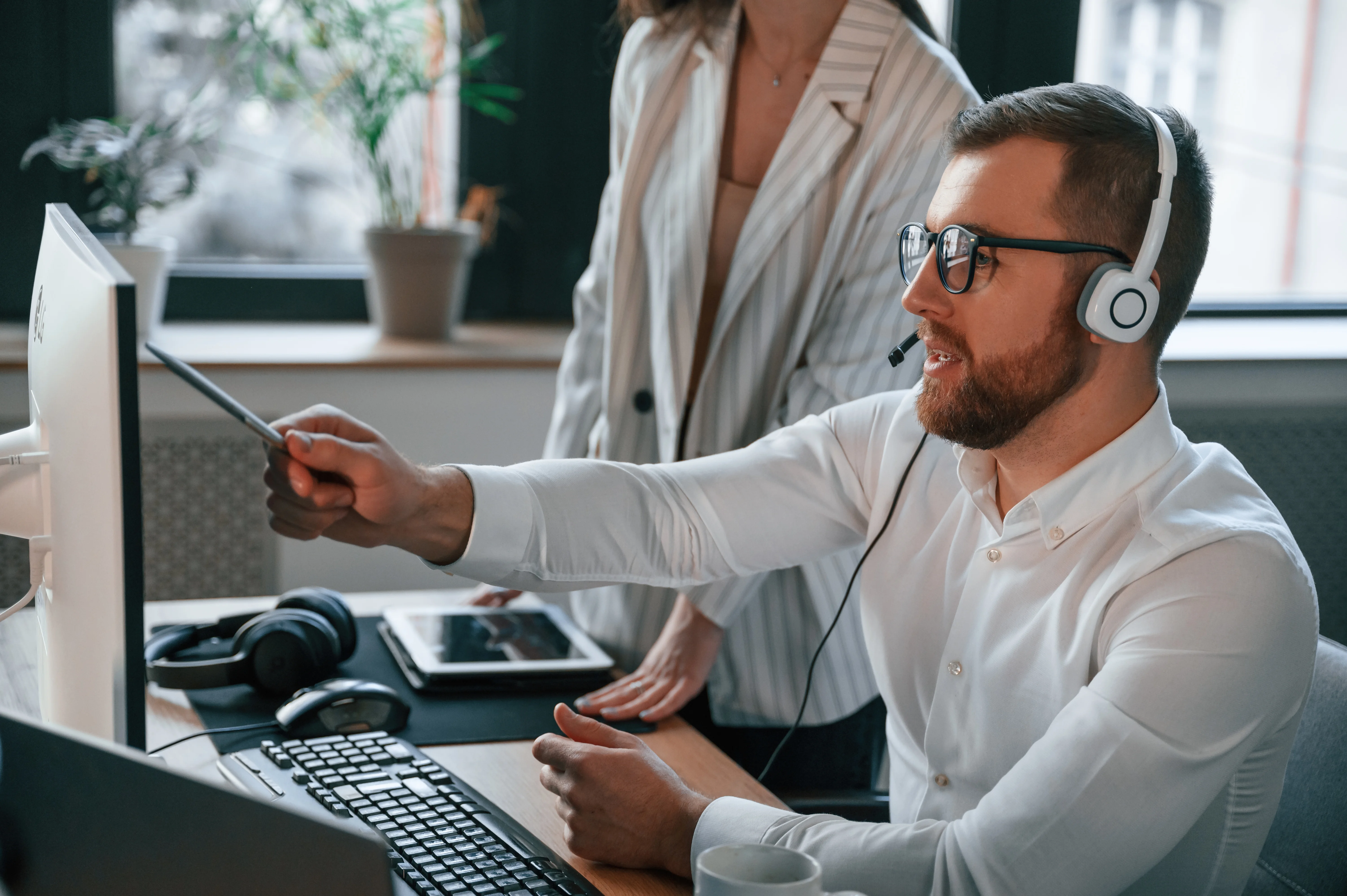 A man sitting at his desk wearing a headset pointing at his computer while a supervisor is watching him.