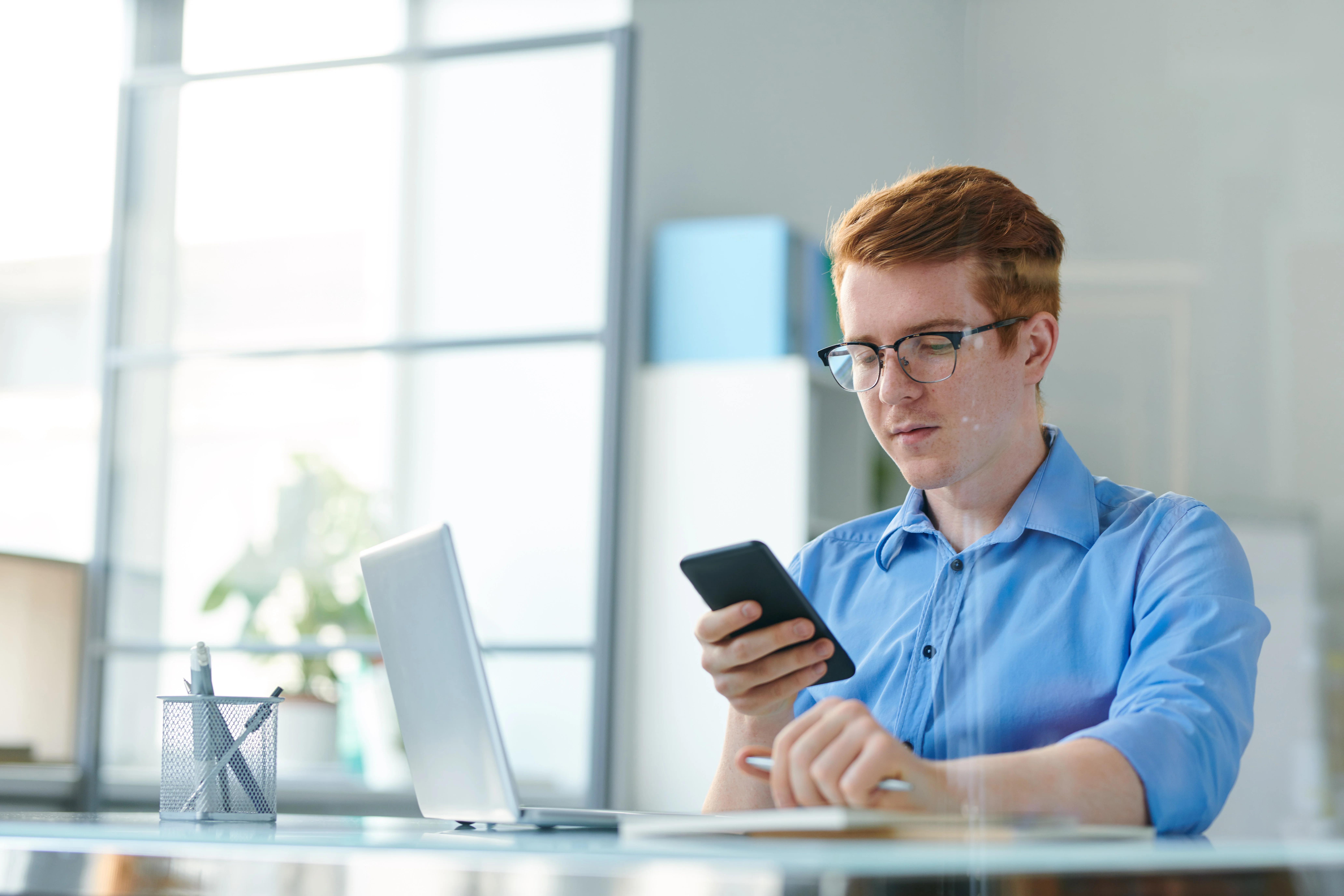 A man sitting at his desk looking at his phone. He is holding a pen and had a computer in front of him.