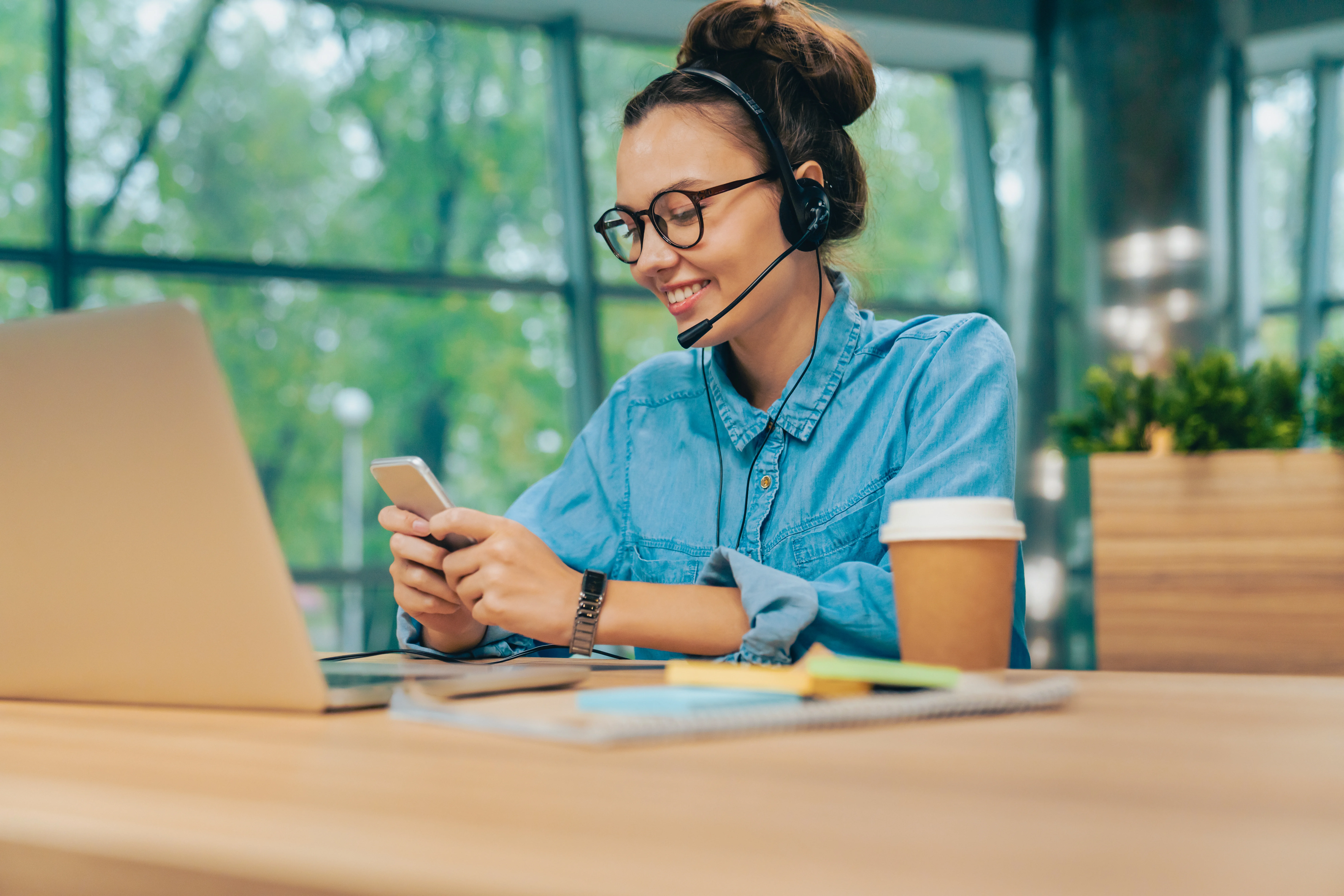 A woman wearing a headset texting on her phone. She has a computer in front of her.