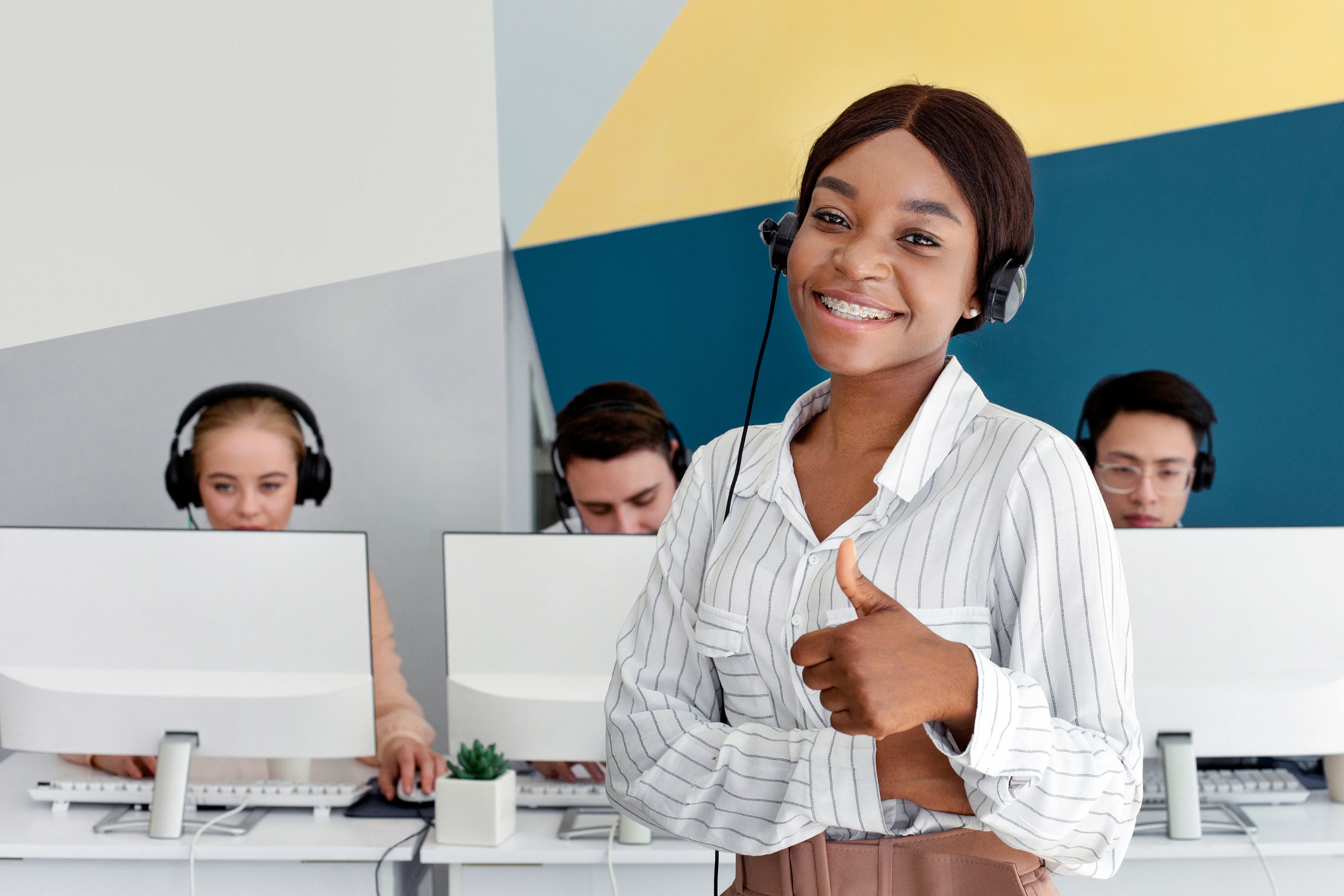 A woman giving a thumbs up while wearing a headset. There are three agents behind her on computers with headsets on.