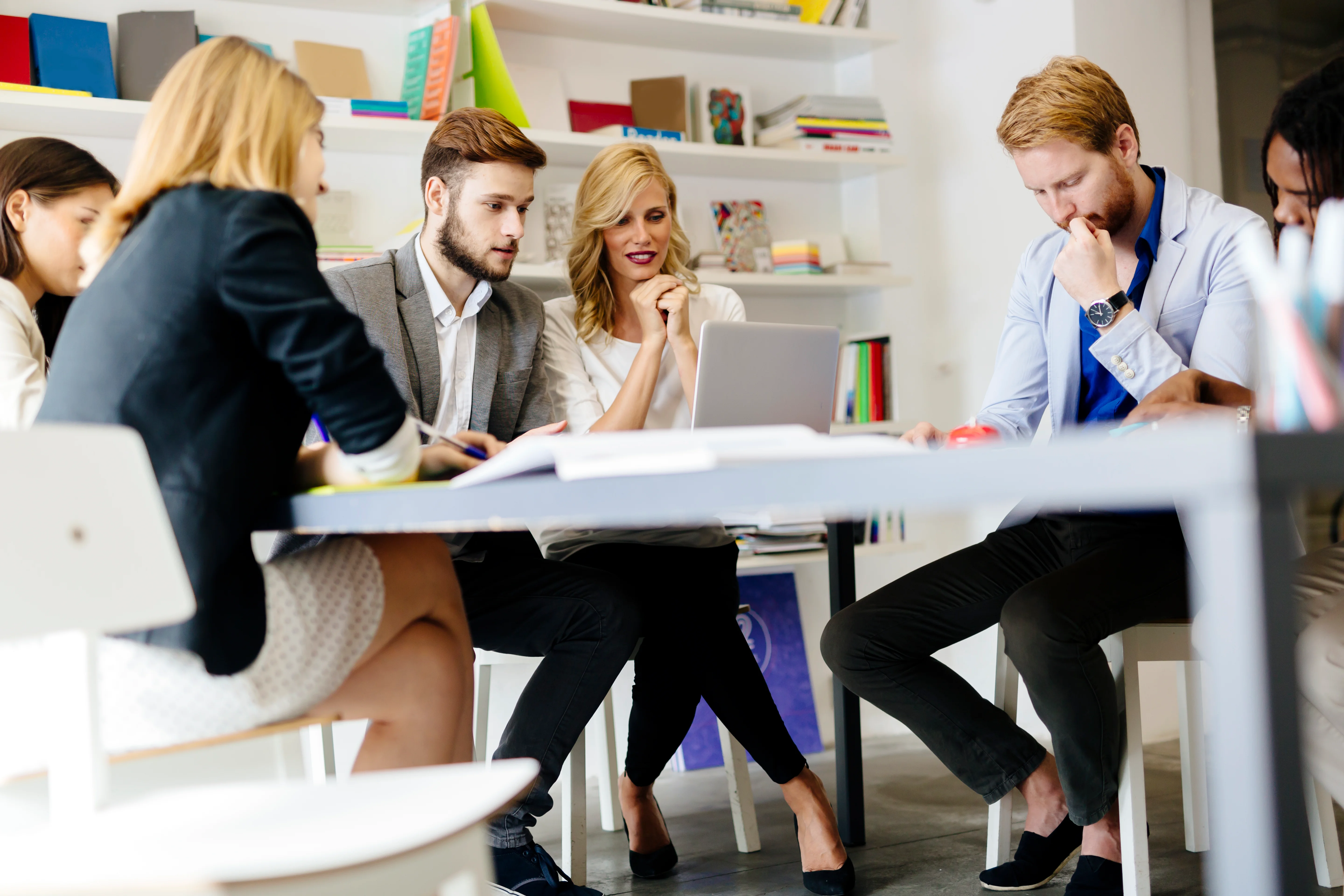 A group of people deciding on something in a conference room. A few of them are looking at a laptop on the table while the rest are looking at the items on the table in front of them.