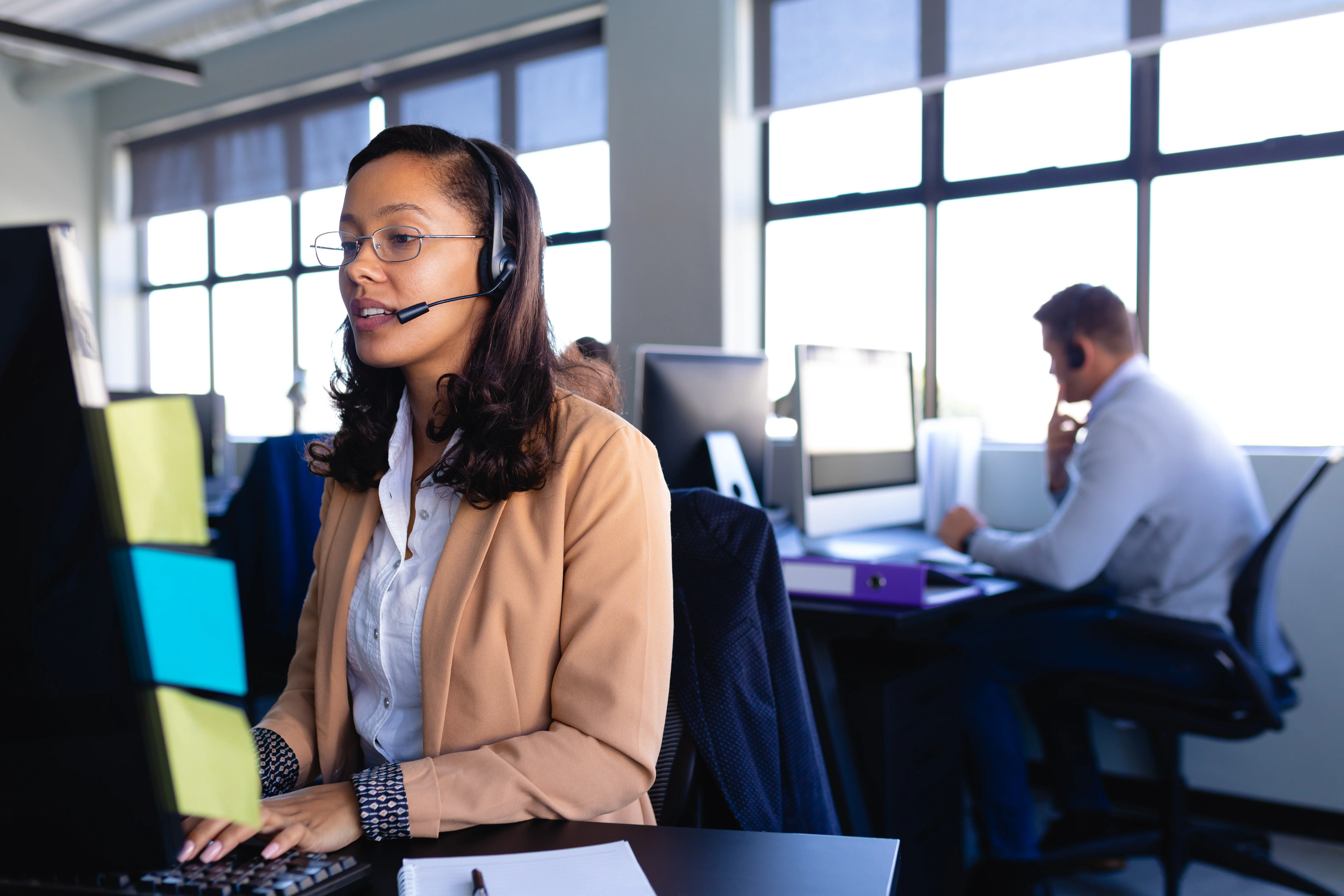 A woman focused on her task at her computer. She is wearing a headset, and her hands are on the keyboard. There is a man in the background contemplating what to say next.