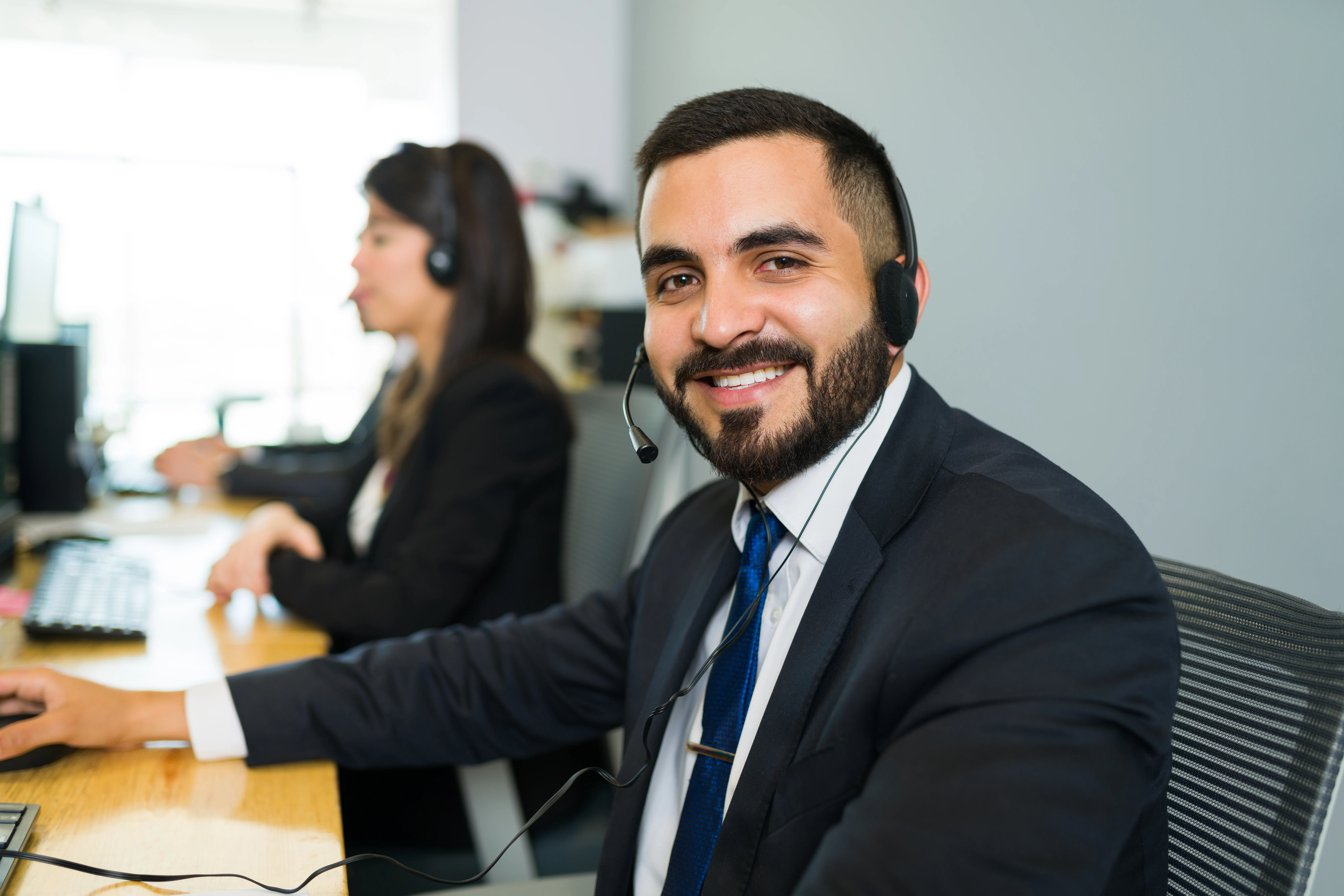 A man wearing a suit and a headset smiling at the camera. There is a coworker next to him.