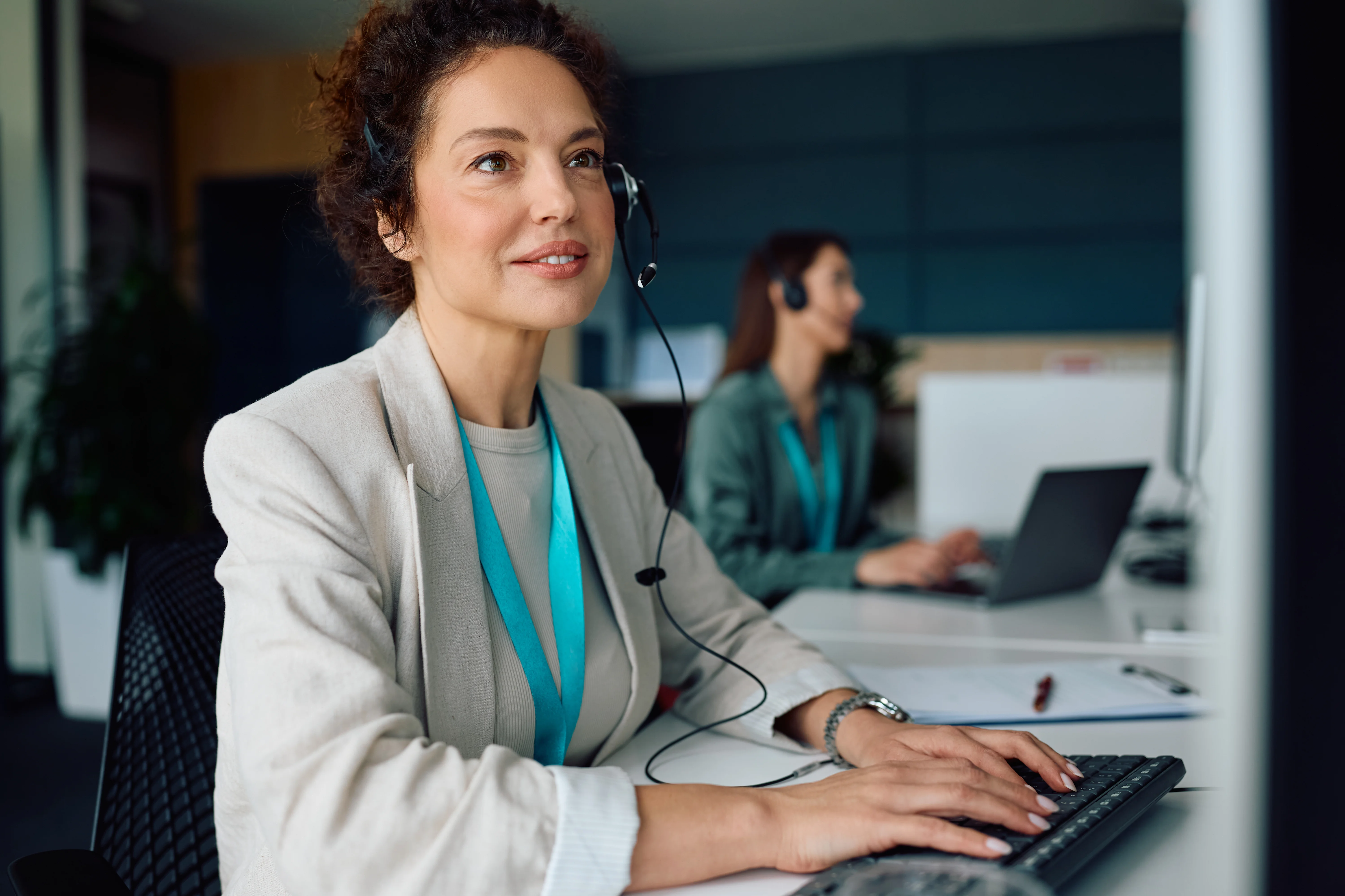 A woman sitting at her computer wearing a light blue lanyard. She's wearing a headset and looking up at the computer screen.