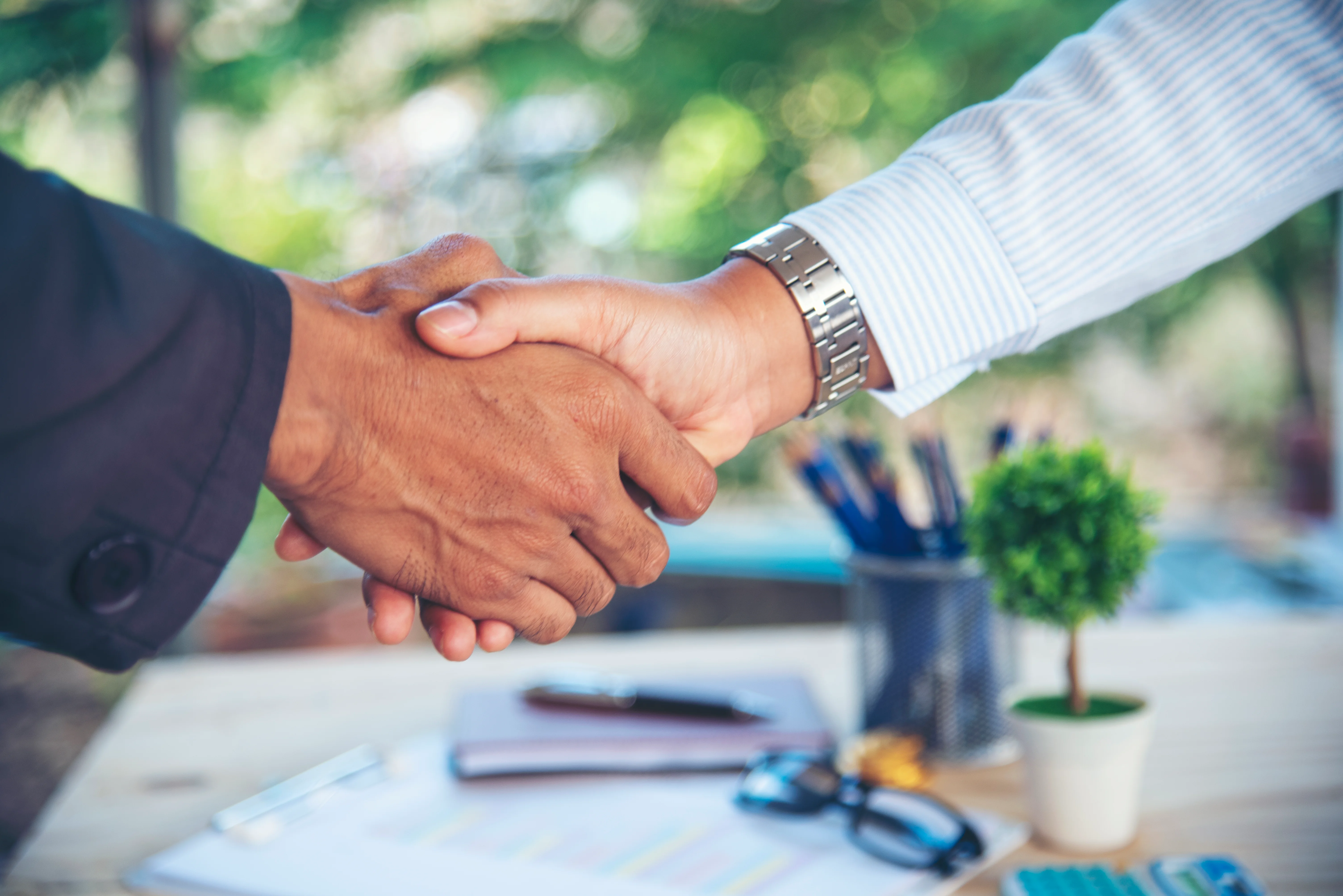 Handshake between two people. There is a notebook, pens, glasses, and a clipboard with papers attached to it on the table.