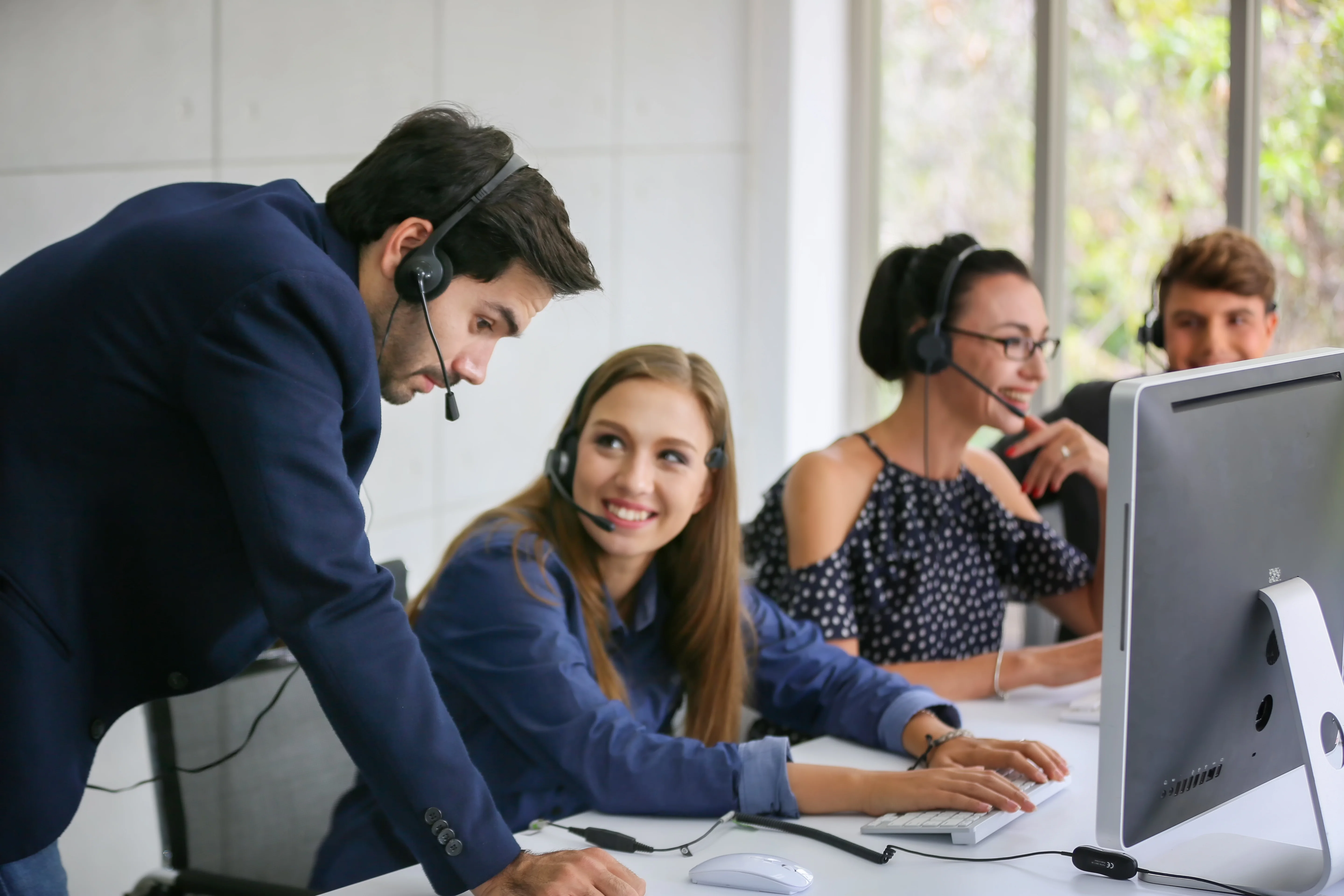 A group of call agents sitting at a communal desk. One woman is getting trained by a man who is leaning on the desk.
